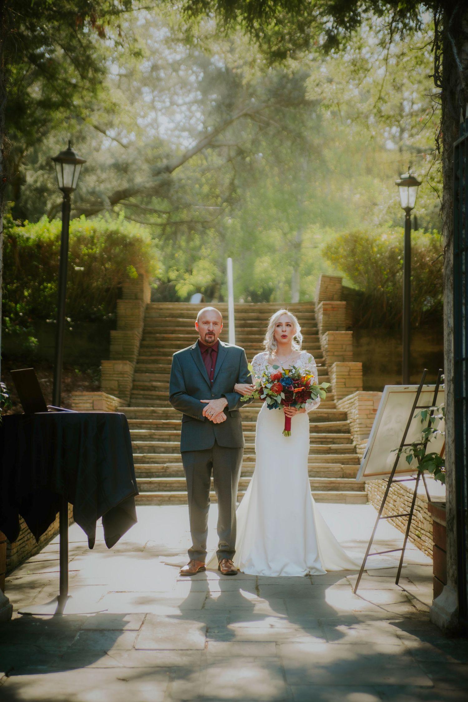 Bride in white gown holding colorful bouquet walks down outdoor aisle with escort amid lush greenery and stone steps.