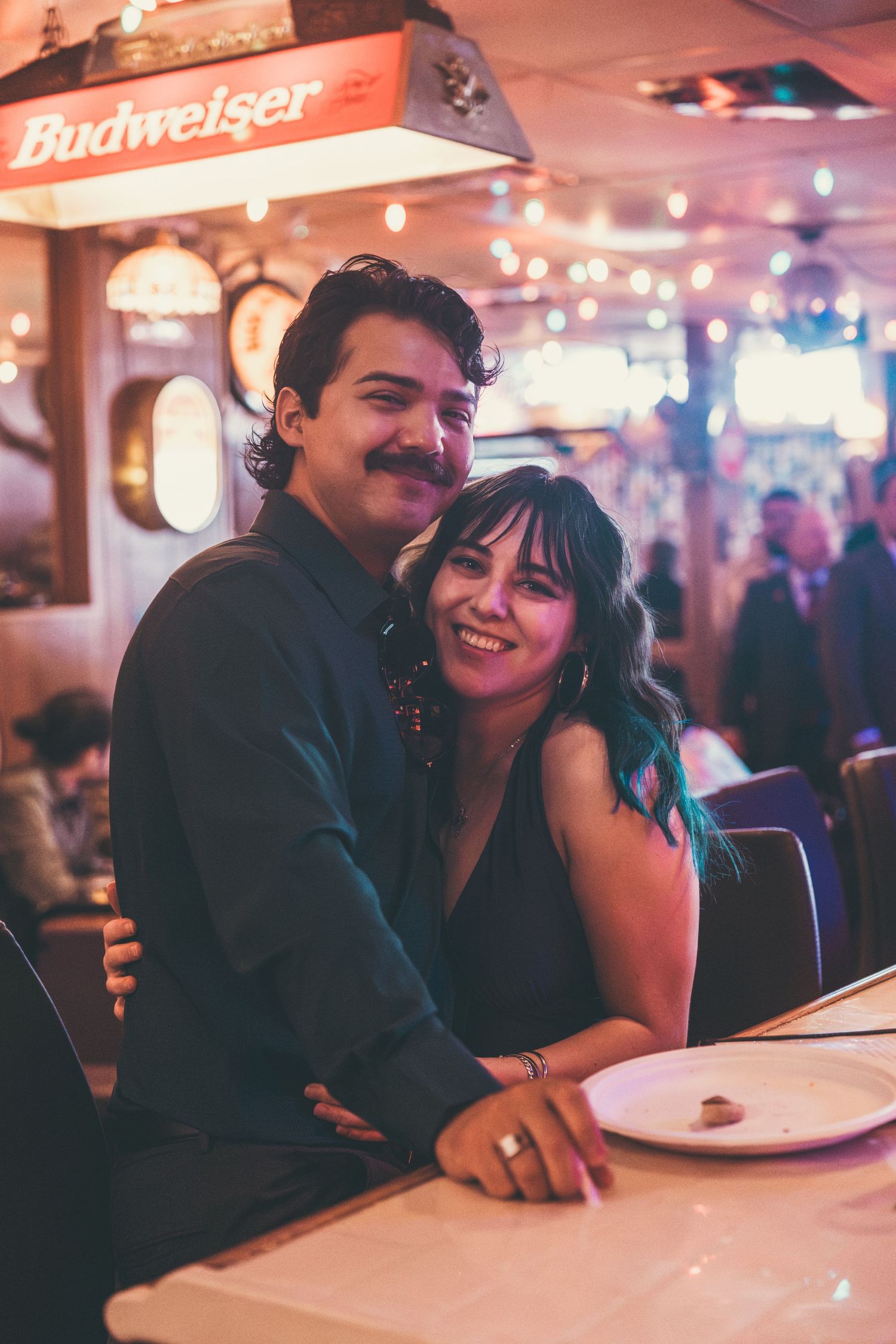Couple smiling together at a Budweiser-lit diner booth, woman wearing dark dress, man with mustache hugging her.