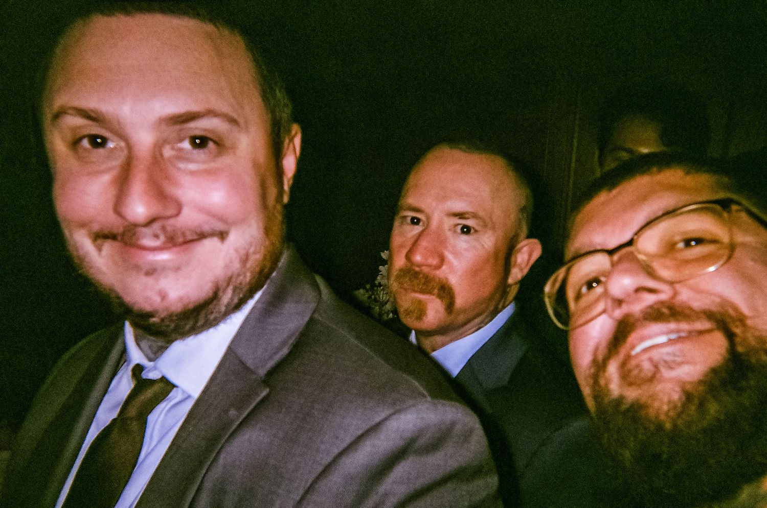 Three men in suits smiling for a selfie photo at a formal event, two with beards, taken in low light.