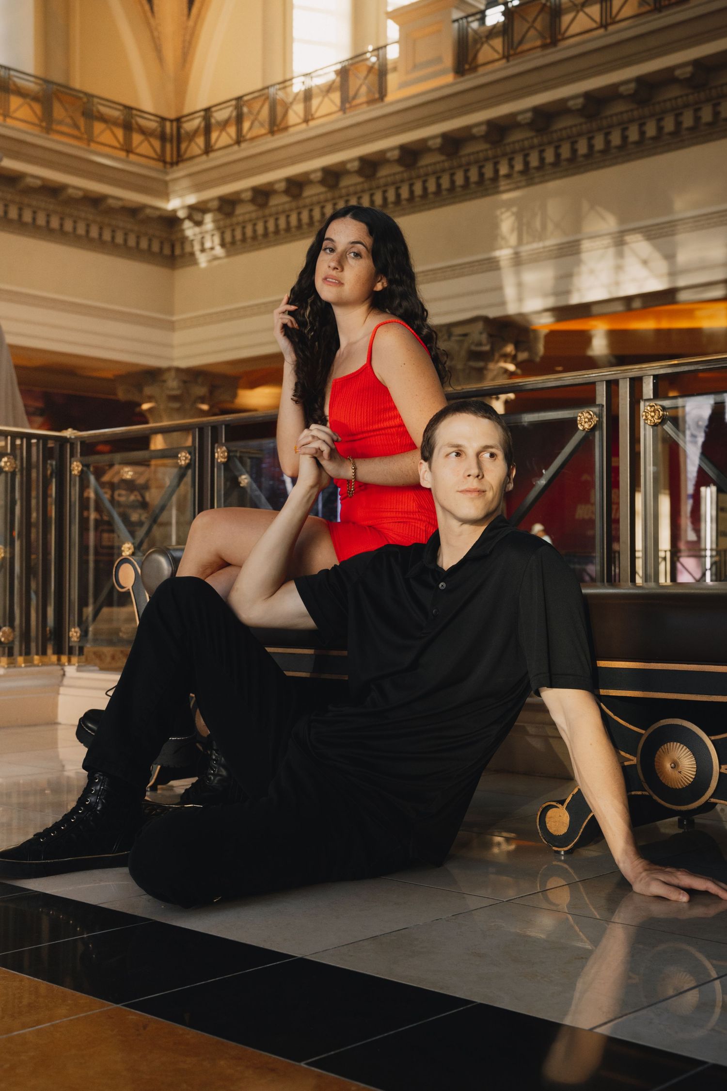 Stylish couple in luxury interior, woman in red dress seated above man in black outfit on marble floor.