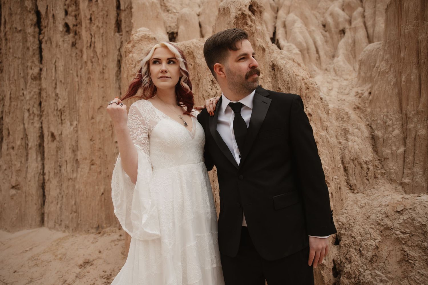 Bride in lace wedding dress and groom in black suit pose before tall sandy rock wall.