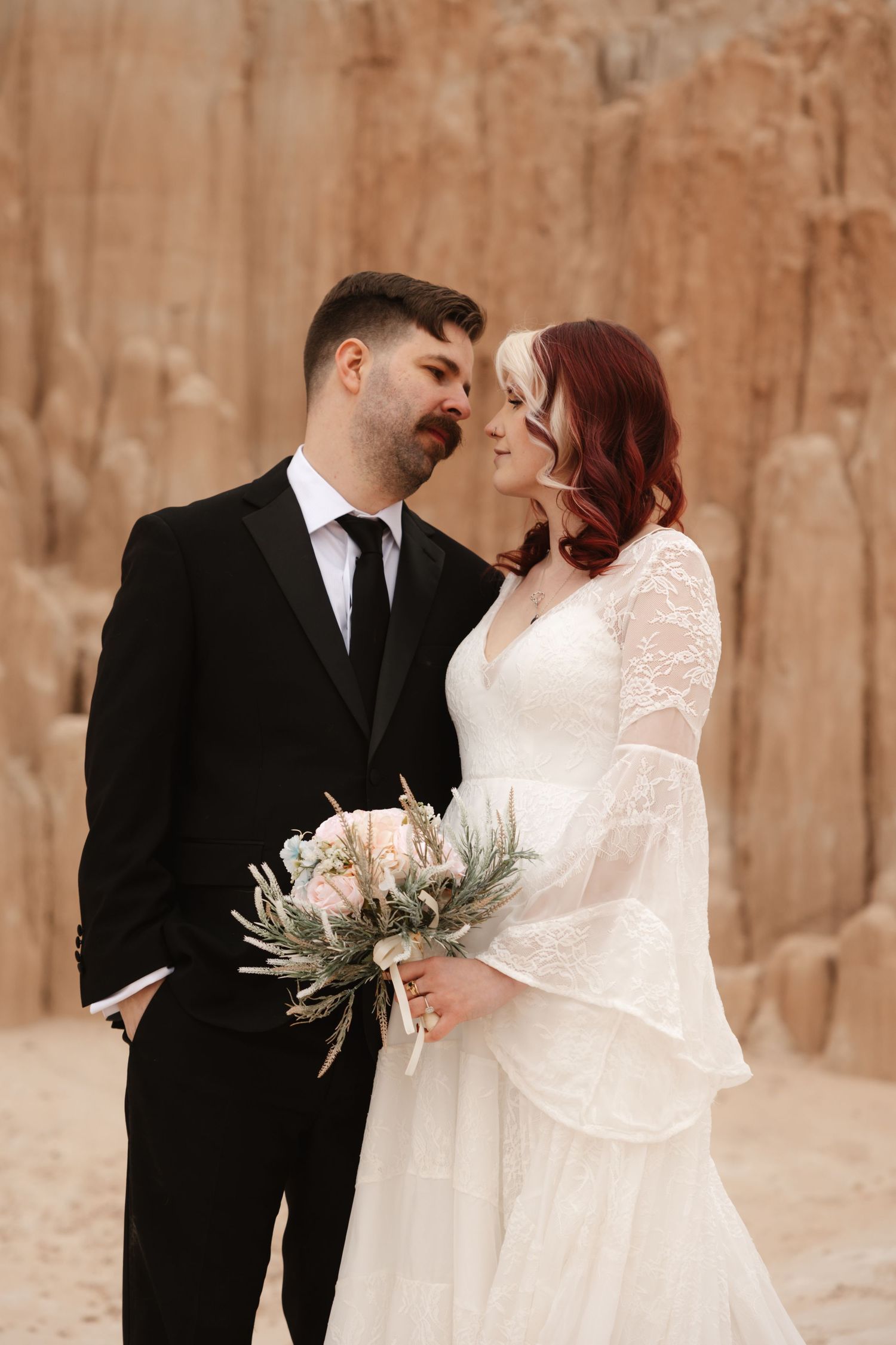 Bride and groom share romantic moment in desert canyon, she holds bouquet in white lace gown, he wears black tuxedo.