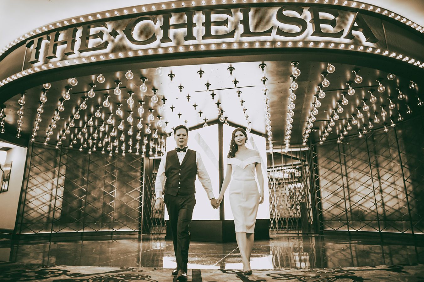 Couple holding hands walking in front of The Chelsea venue entrance with marquee lights in sepia tone.