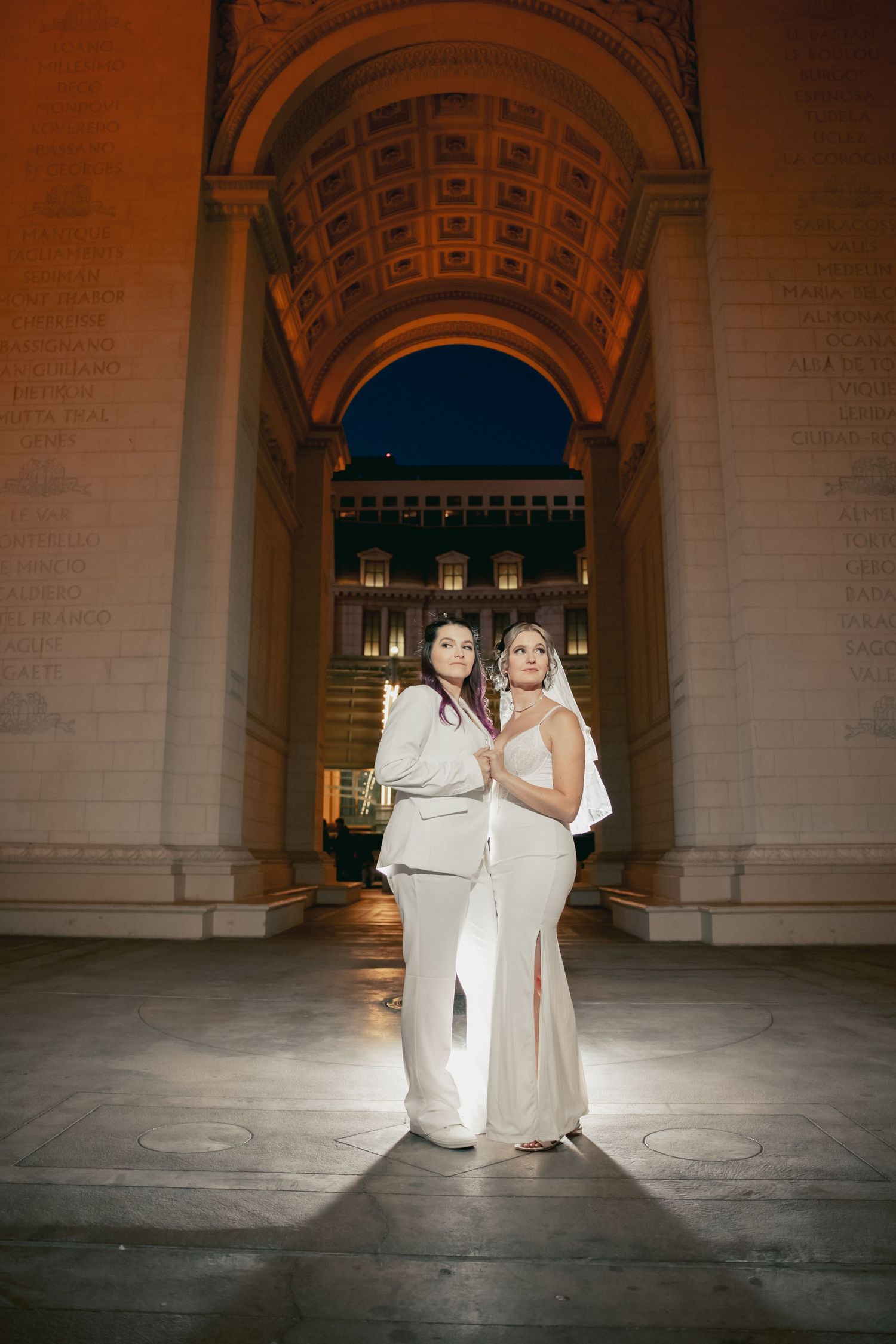 Two brides in white attire pose under an illuminated grand arch at night.