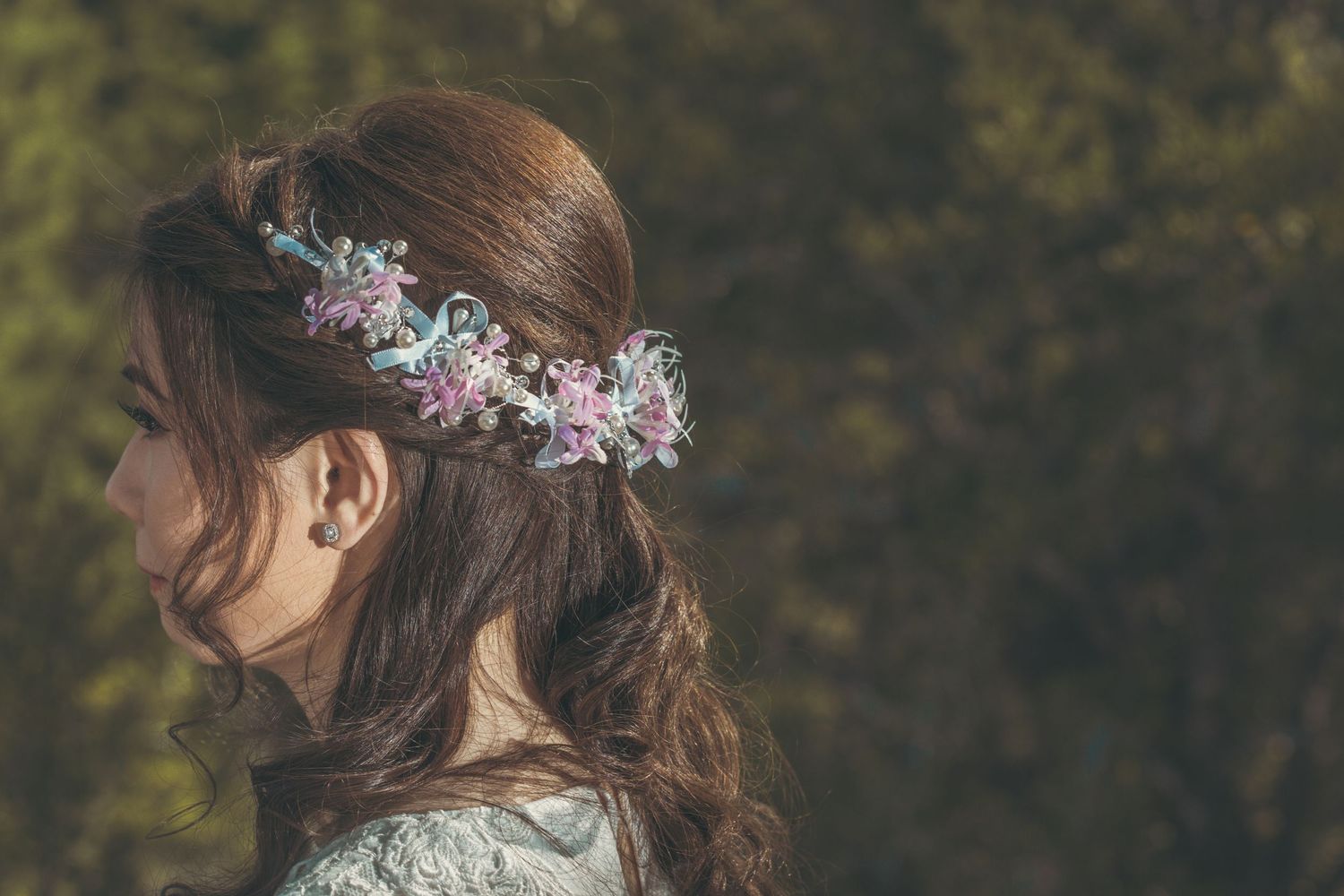 Young woman with long brown hair wearing a delicate floral crown of pink and blue flowers, photographed outdoors.