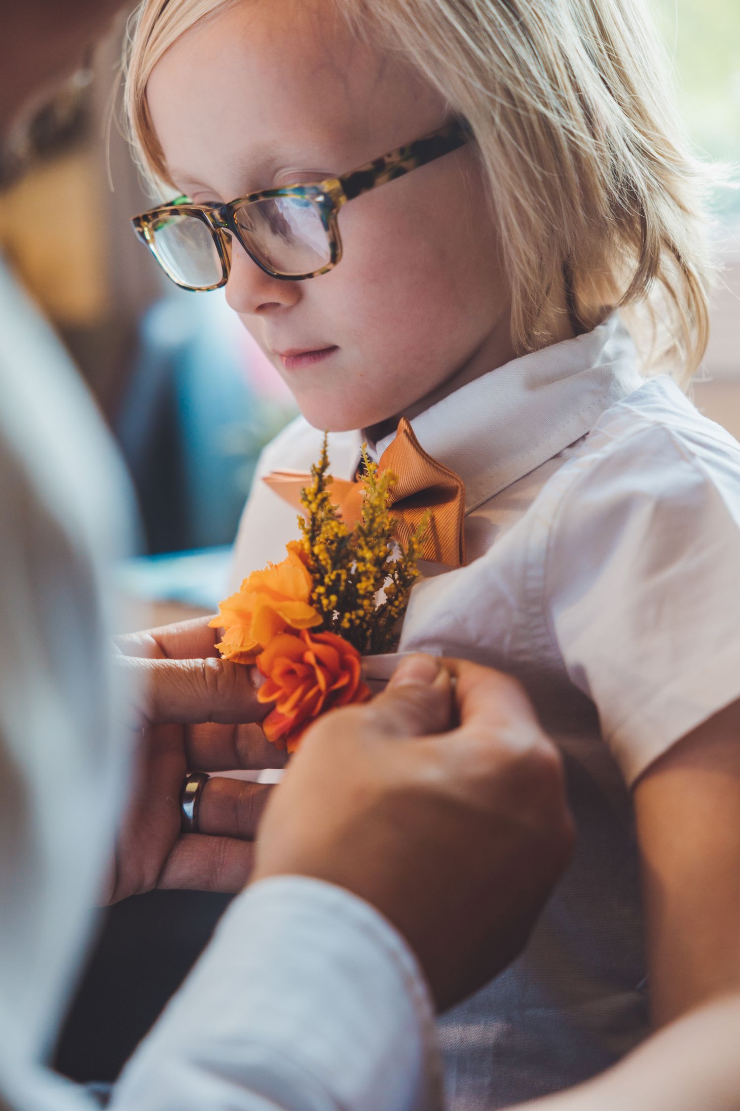 Blonde child with glasses having an orange boutonniere pinned to white shirt at a wedding.