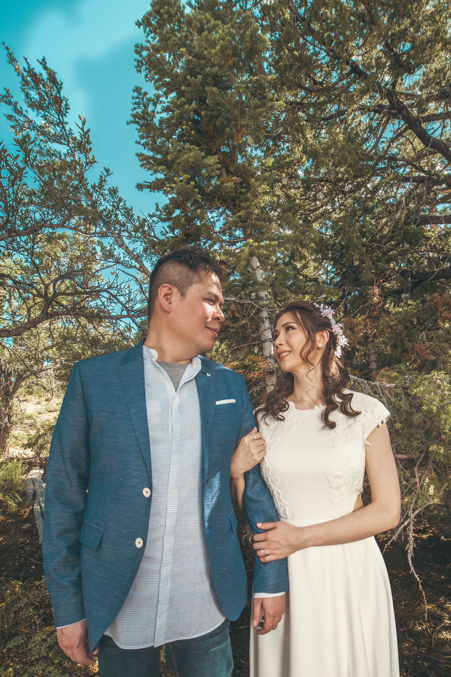 Couple in wedding attire smiling at each other outdoors among pine trees under a blue sky.