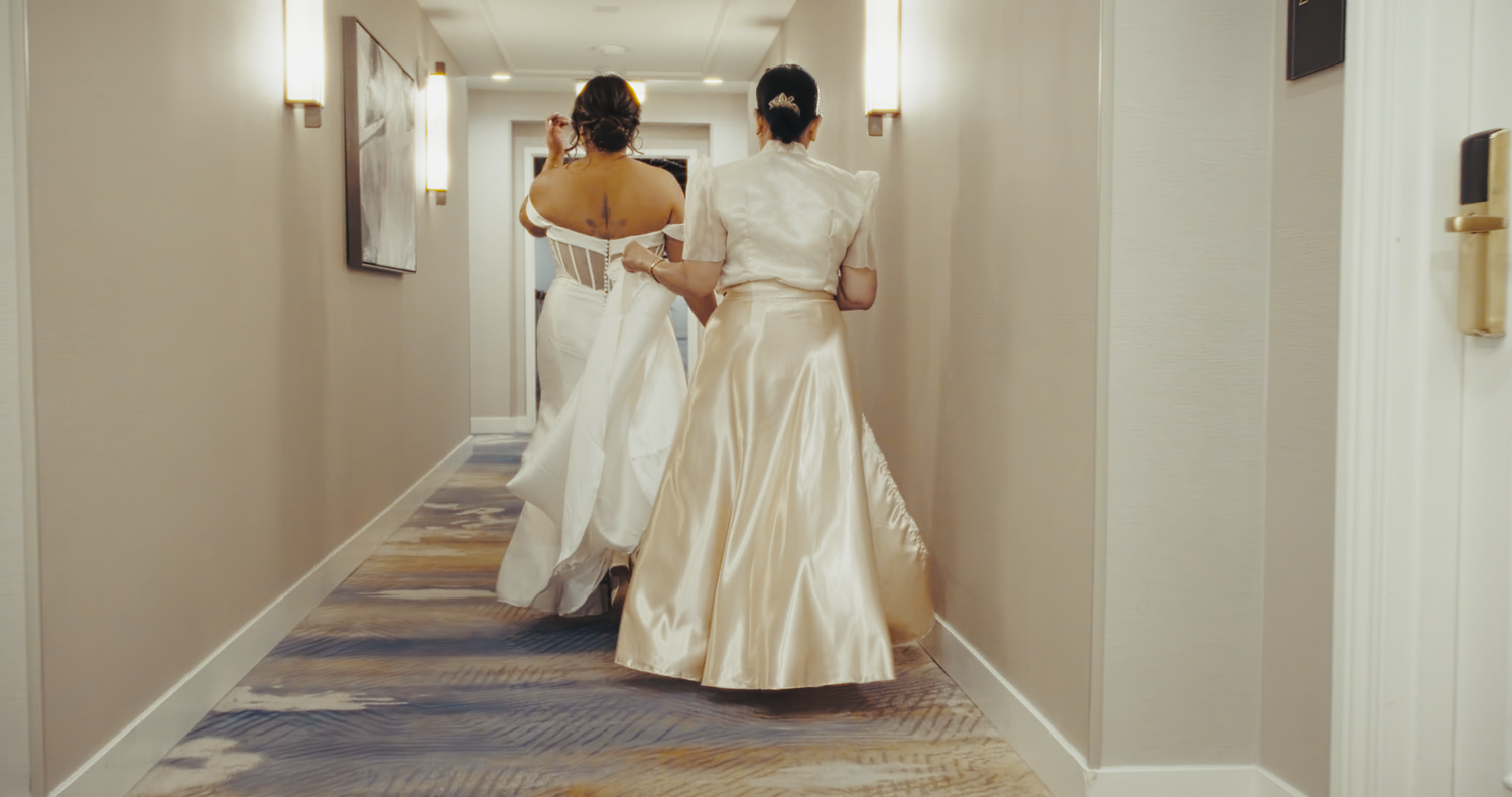 Two women in white bridal gowns walk down a hotel corridor from behind.