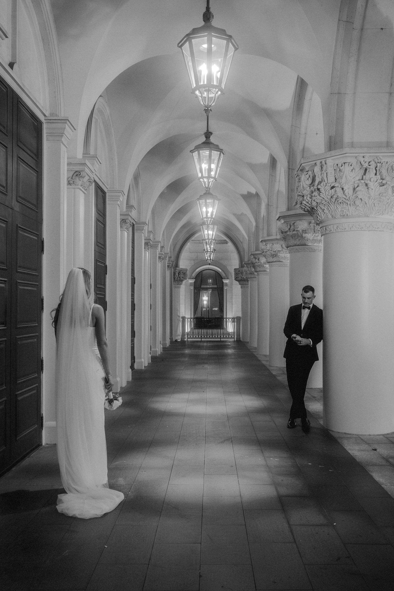 Black and white photo of bride and groom in elegant arched corridor with hanging lanterns.