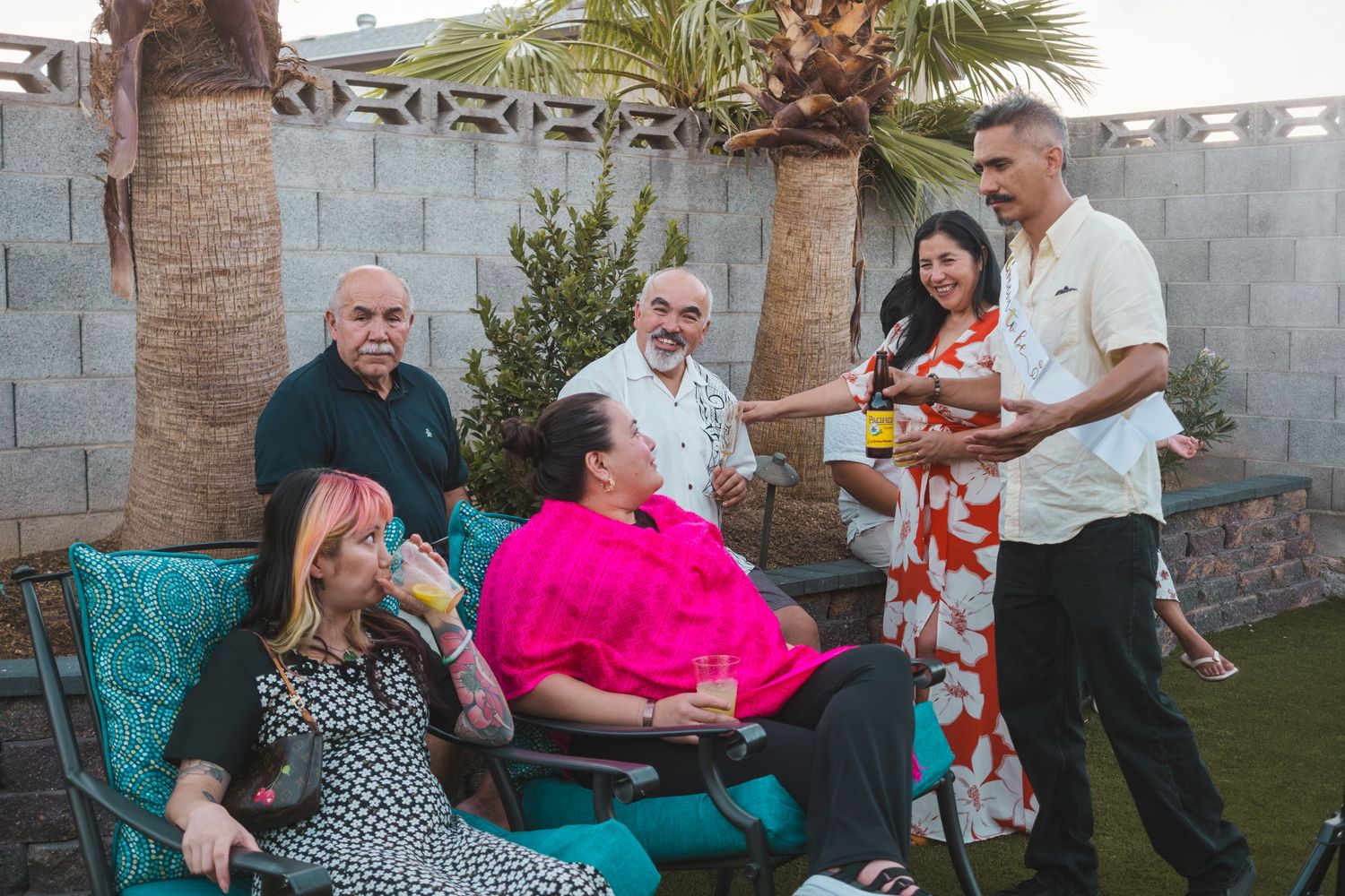 Group of friends and family gathered outdoors at a backyard party, socializing and enjoying drinks near palm trees.