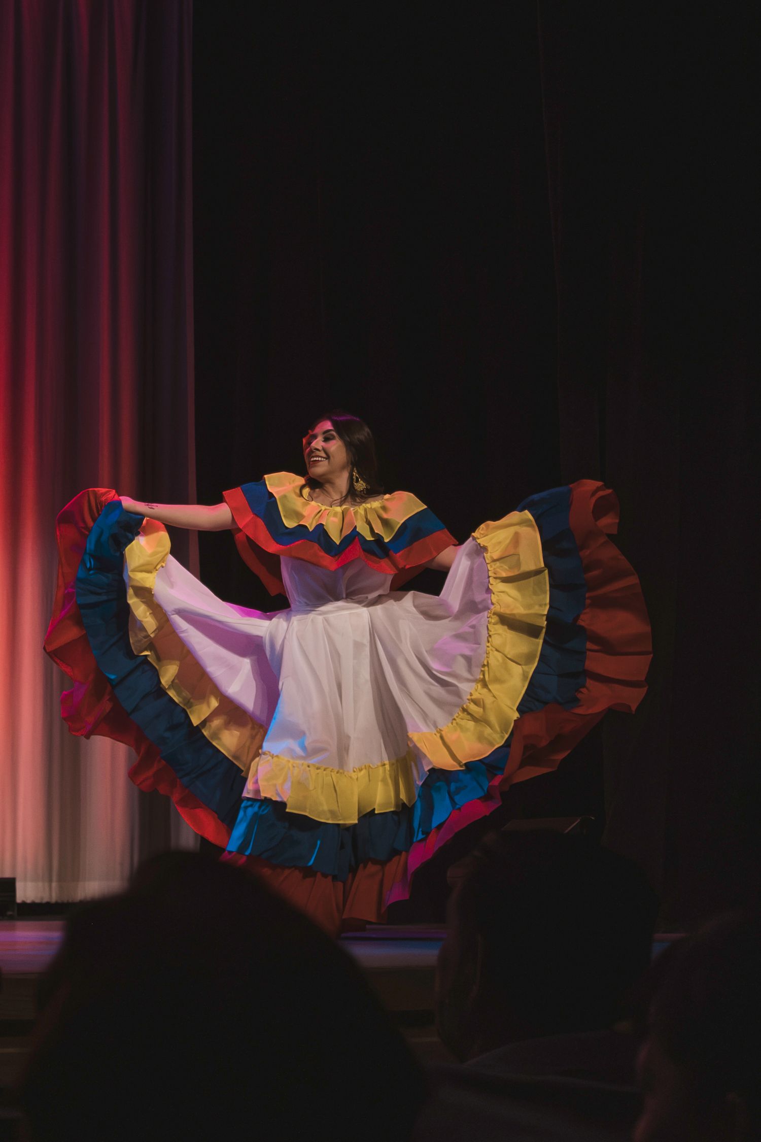 Colorful traditional Latin dancer performs on stage in vibrant ruffled dress with red, blue, and yellow layers.