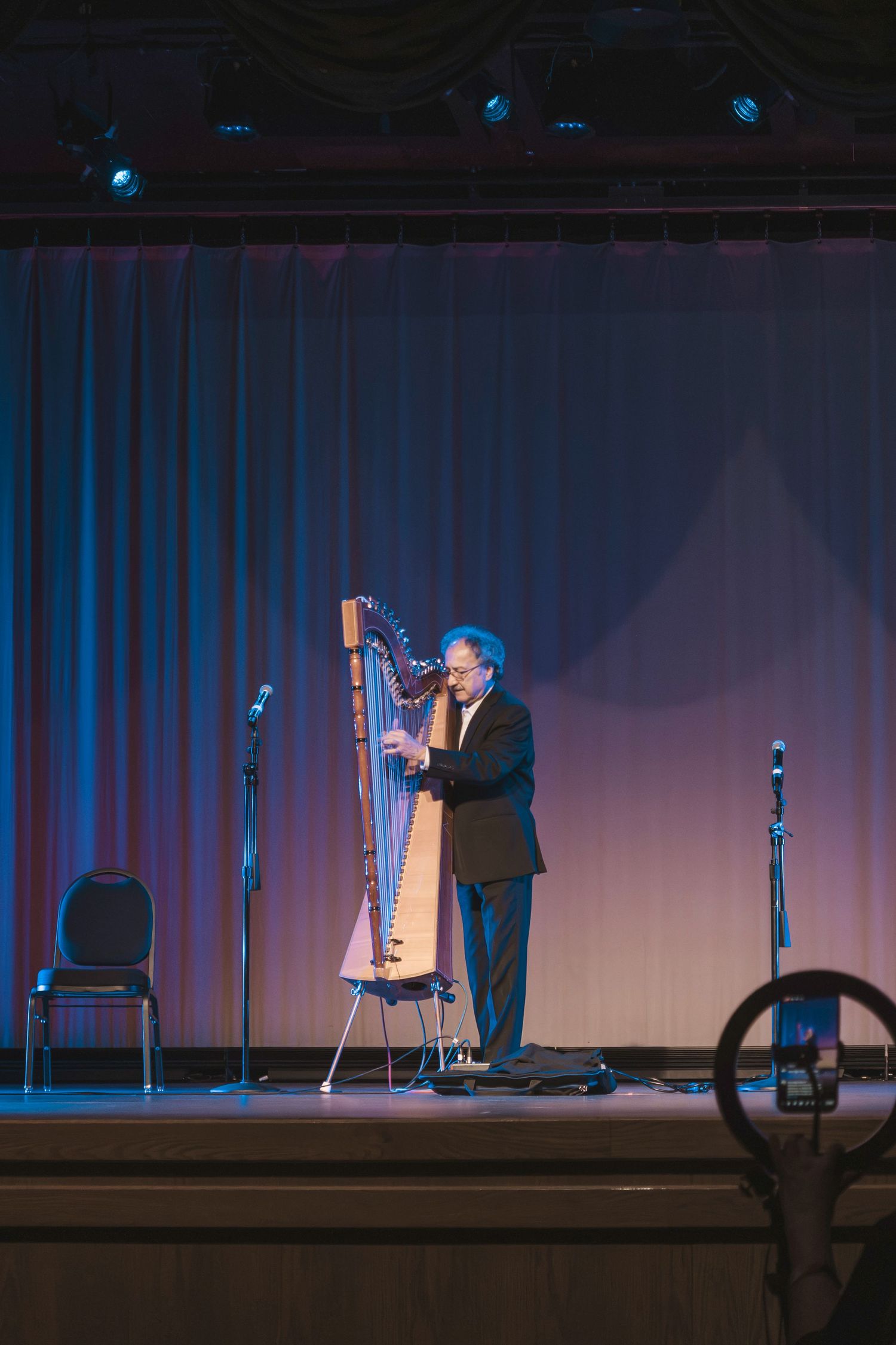 A man in a black suit plays a large harp on a stage with blue and pink lighting and a curtain backdrop.