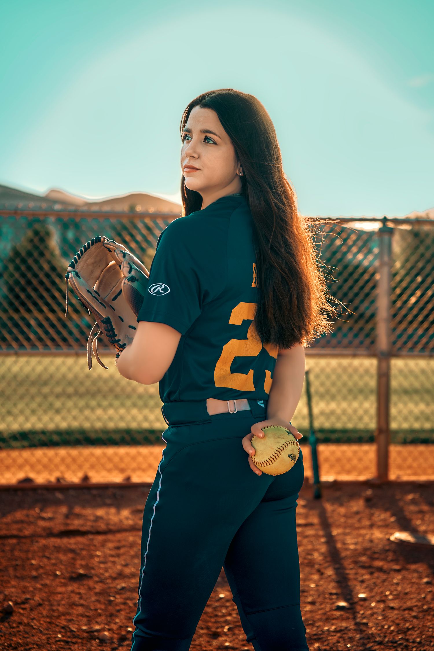 Female softball player in teal uniform holding glove and ball on dirt field at sunset.