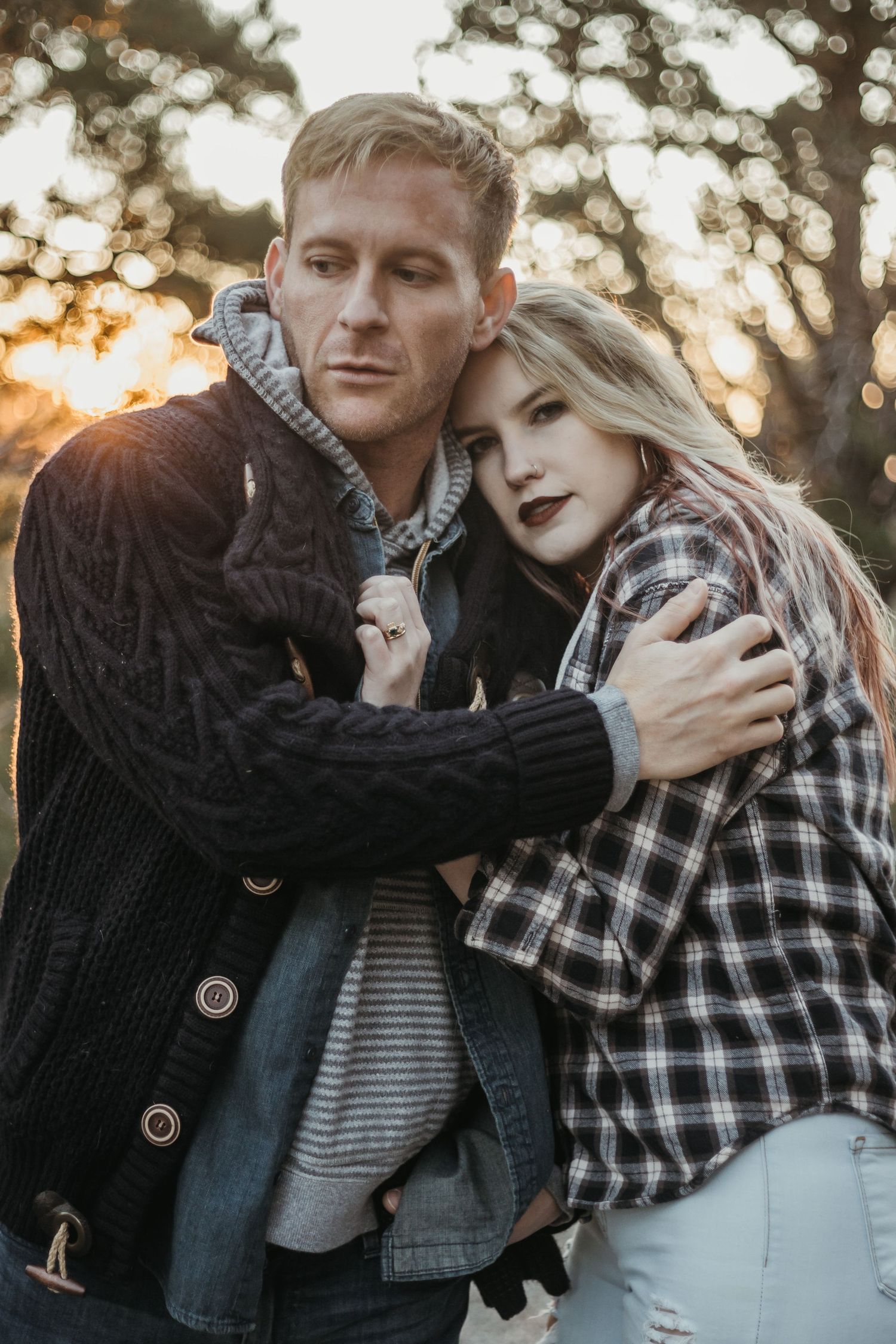 Couple embracing outdoors at golden hour, man in knit sweater hugging woman in plaid shirt among trees.