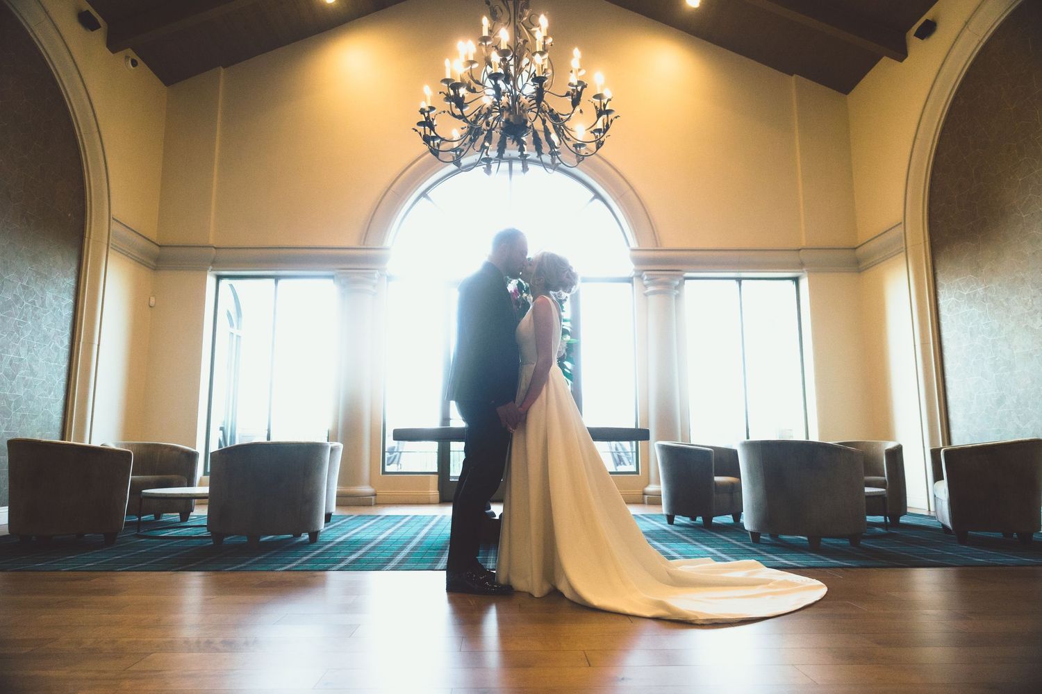Silhouette of a bride and groom kissing beneath a chandelier in a sunlit elegant venue interior.