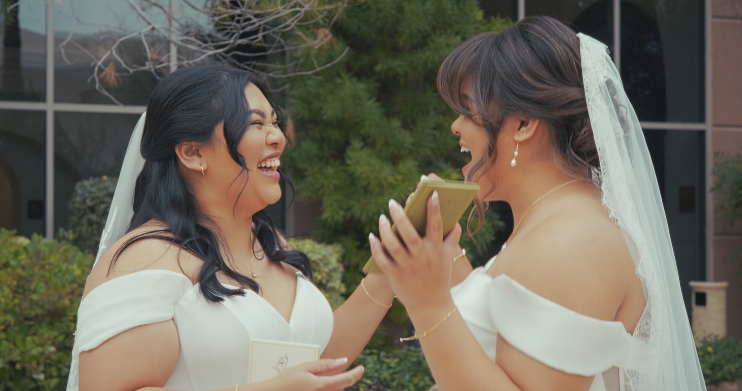 Two brides in white dresses laughing joyfully together outdoors.