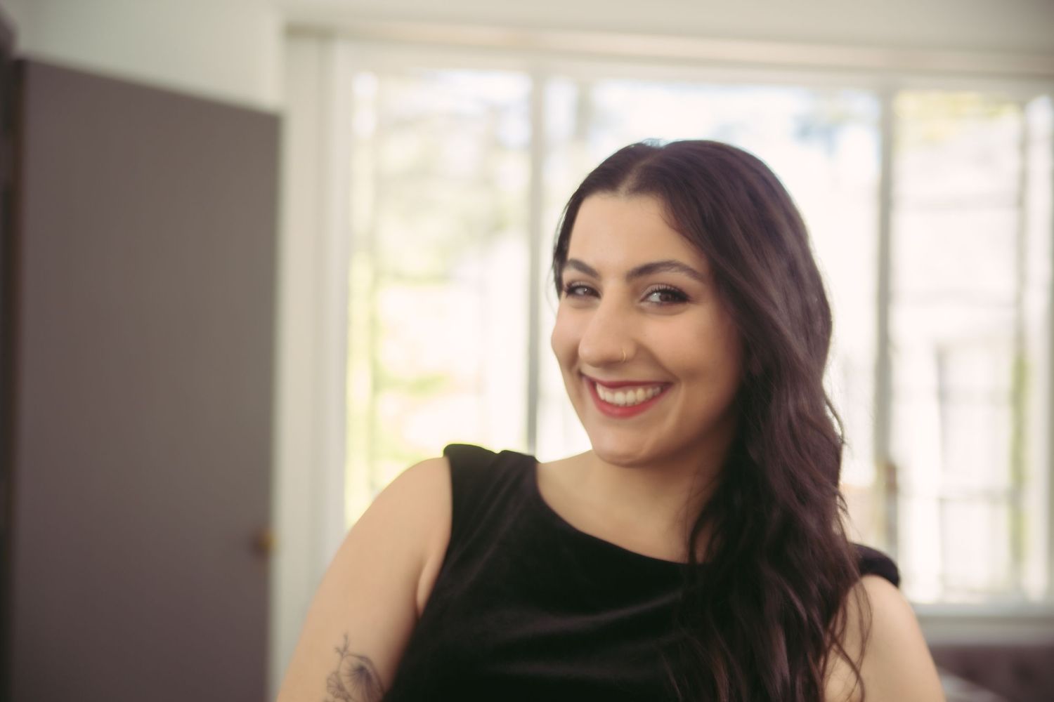 Happy woman with dark hair and tattoo wearing black sleeveless dress smiling indoors.