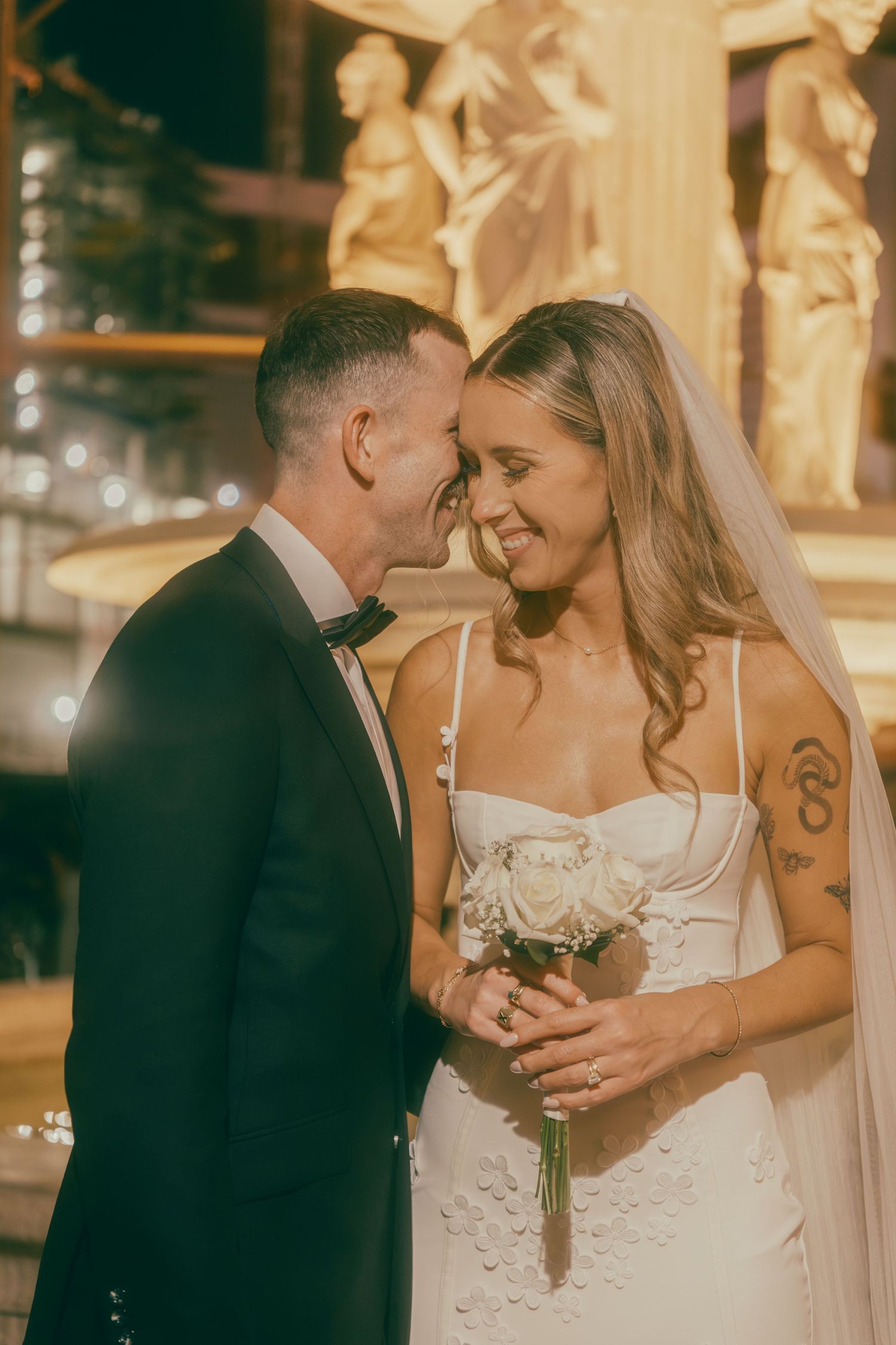 Laughing bride in white veil and groom in tuxedo share intimate moment before golden fountain at night.
