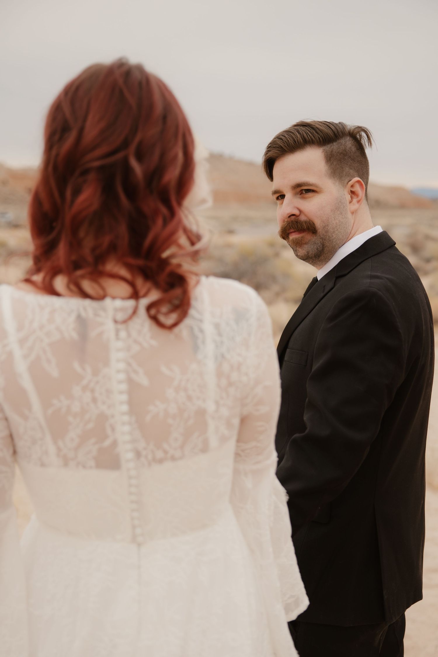 Groom in black suit gazes lovingly at bride in lace wedding dress during desert elopement ceremony.