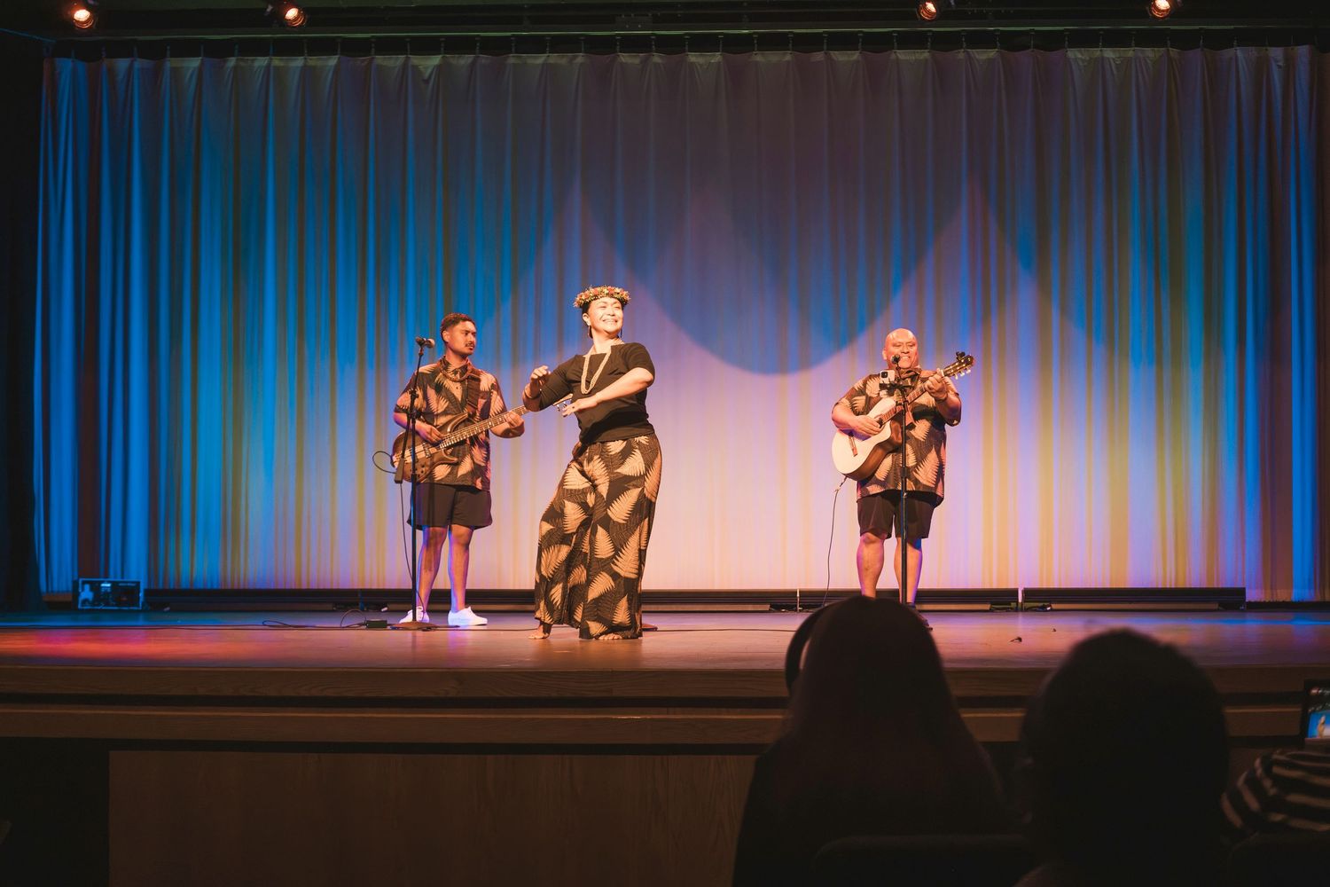 Three performers on stage with blue curtain backdrop, two playing guitars in Hawaiian shirts and one in floral dress.