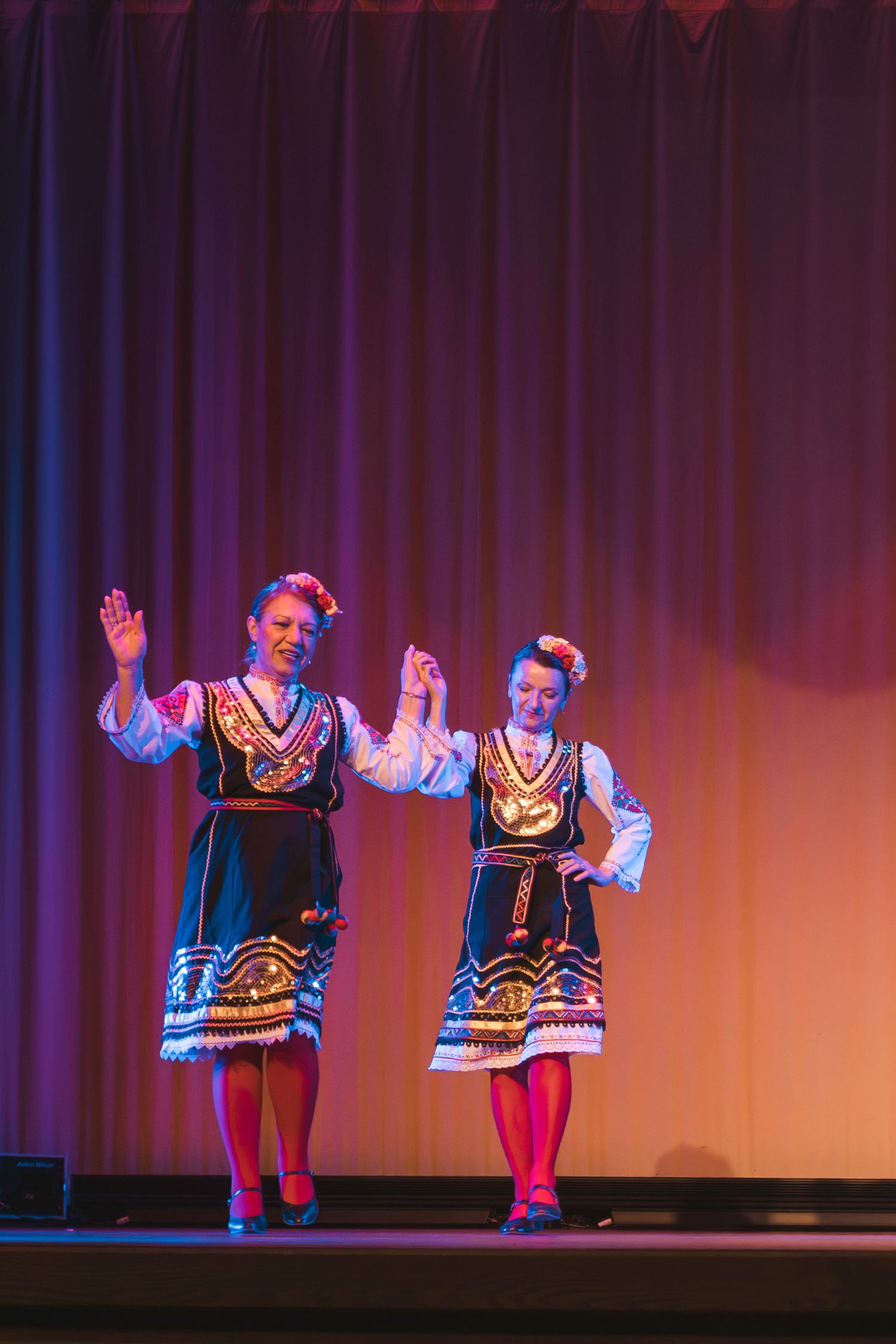 Two women performing traditional folk dance on stage in colorful embroidered costumes with black aprons and floral headpieces.