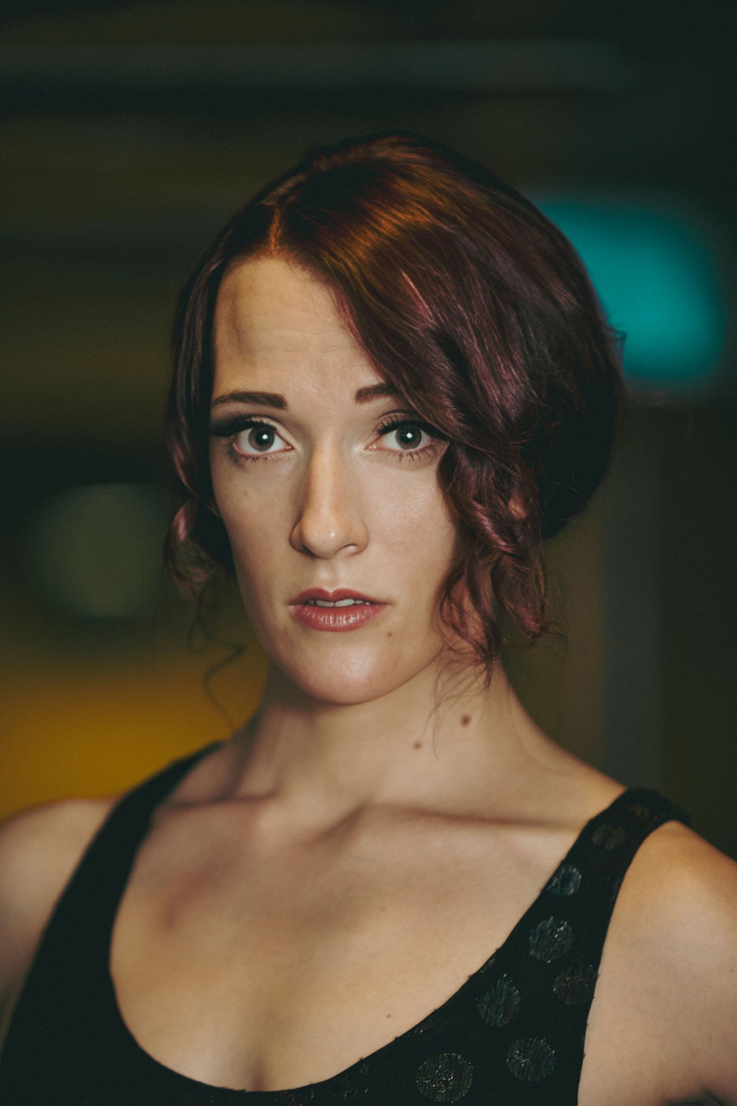 Portrait of a young woman with auburn updo hair, blue eyes, freckles, and a black lace top against a dark background.