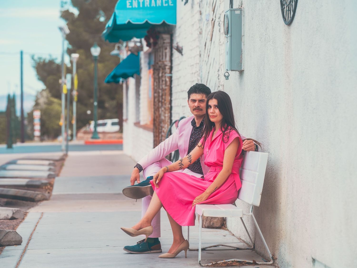 A couple sits on a white bench outside a building with a teal awning on a sunny sidewalk.
