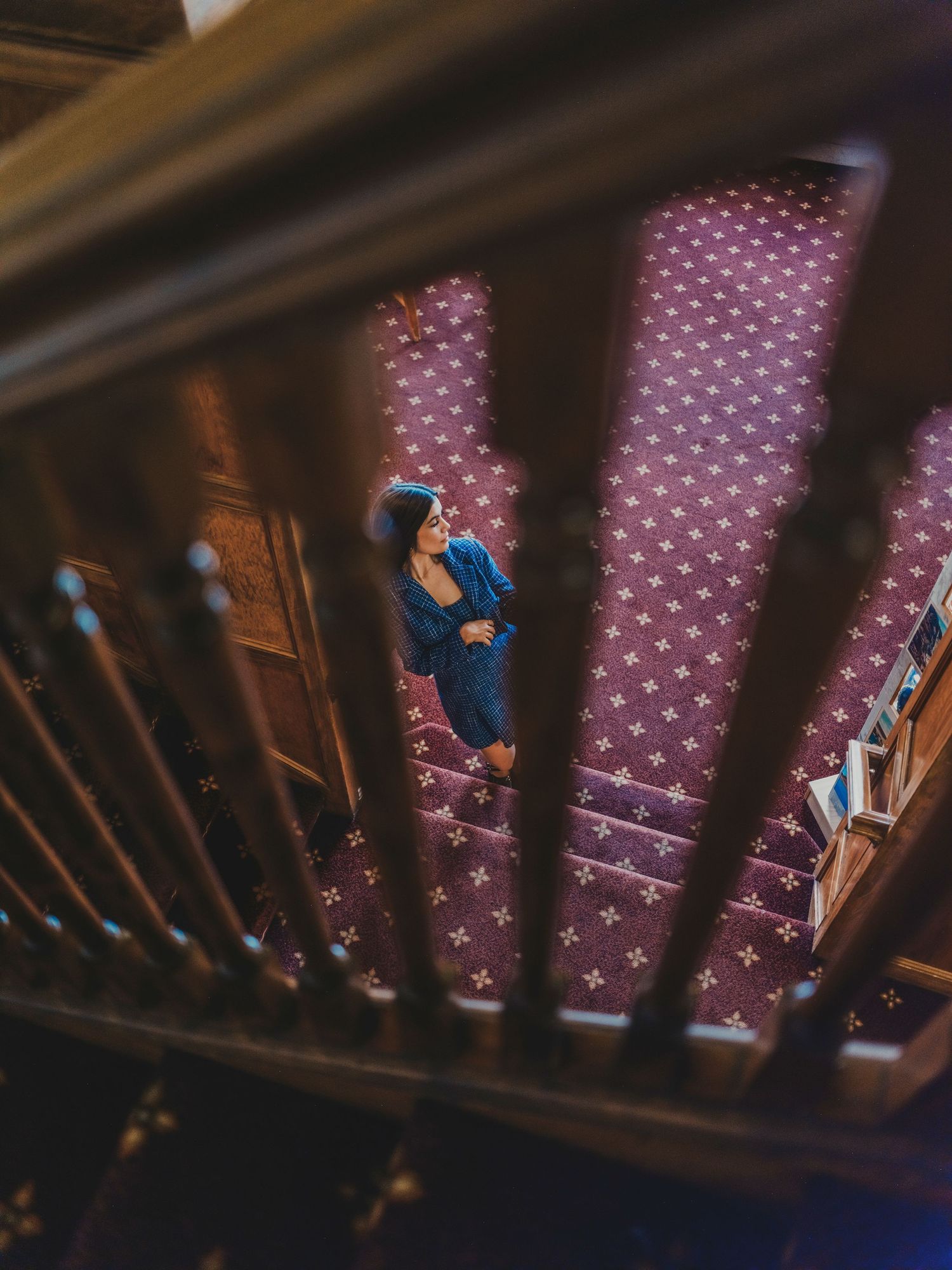 A woman in a blue dress stands on a patterned purple carpet, viewed from above through wooden stair railings.