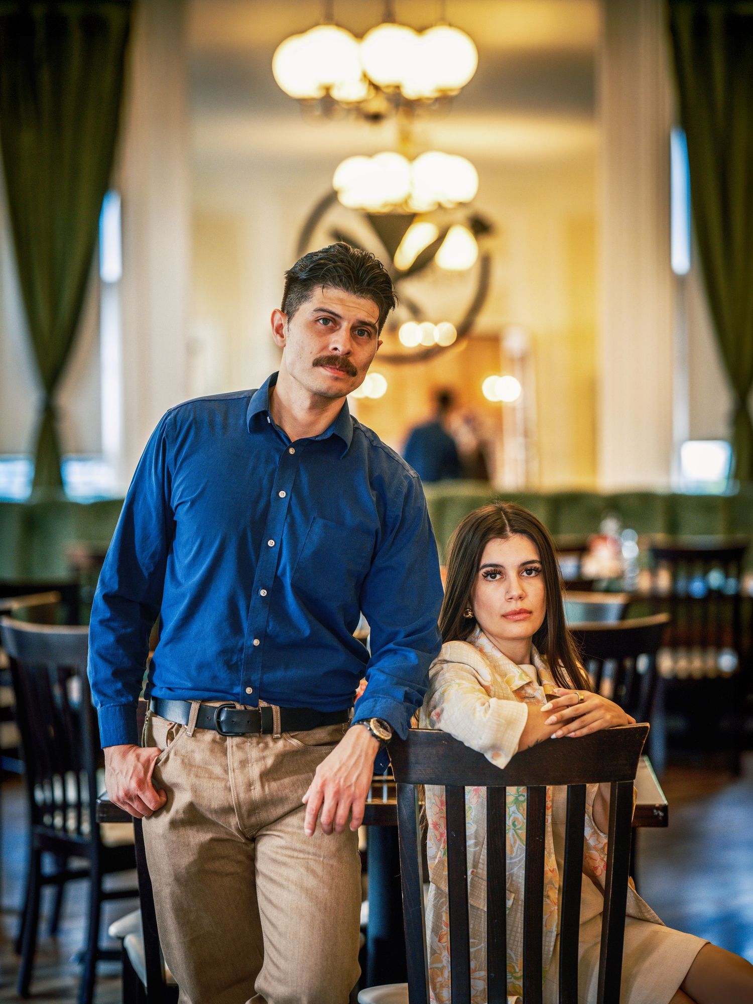 A man in a blue shirt and woman in white pose elegantly in an upscale restaurant with chandeliers.