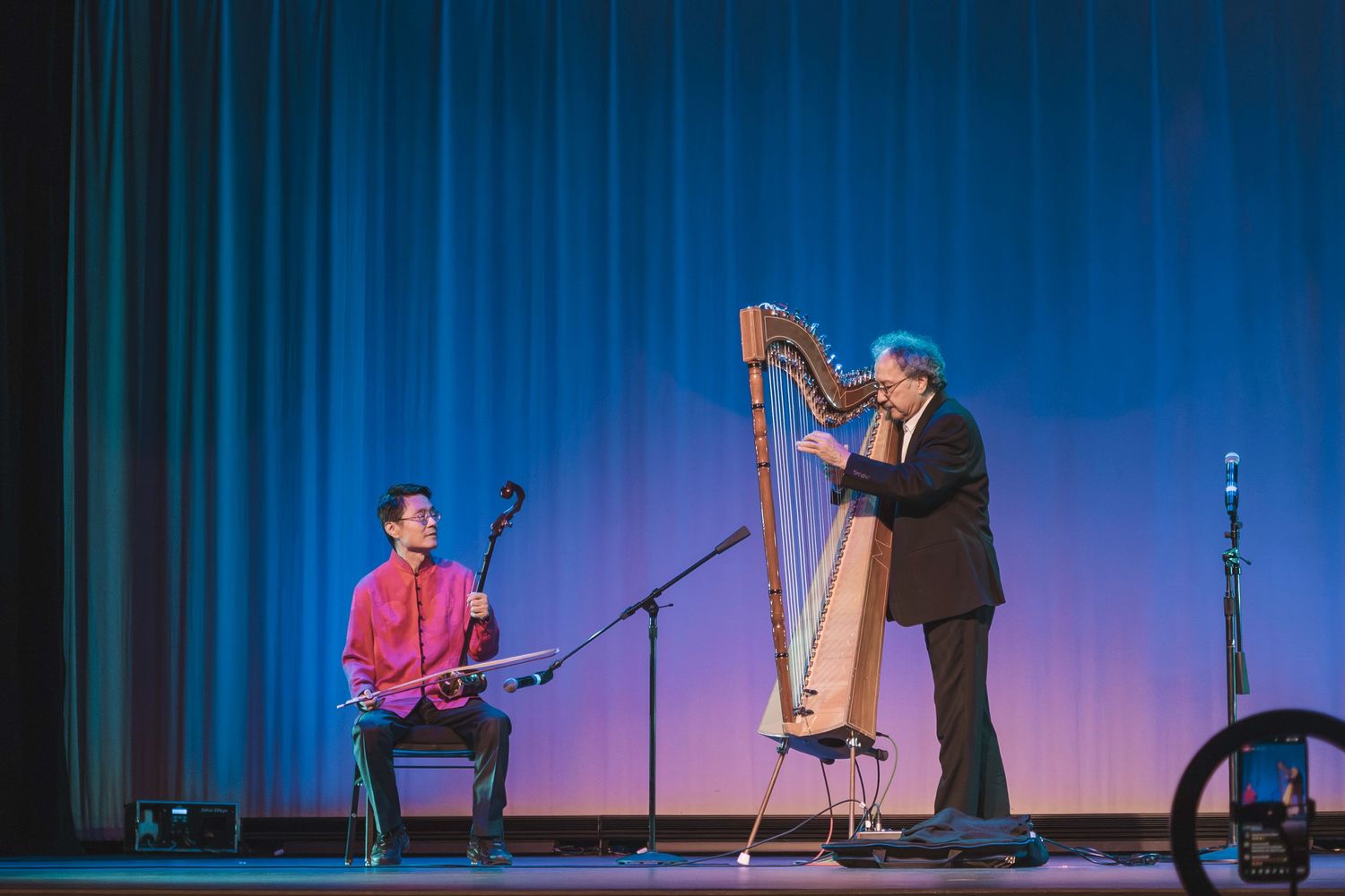 Two musicians perform on stage, one playing guitar and one playing harp, in front of a blue curtain backdrop.