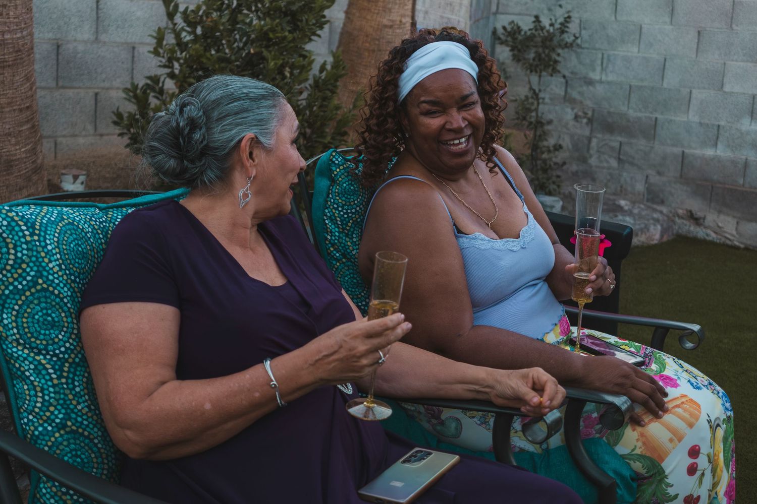 Two women laughing and enjoying wine glasses together while seated outdoors at a casual backyard gathering.