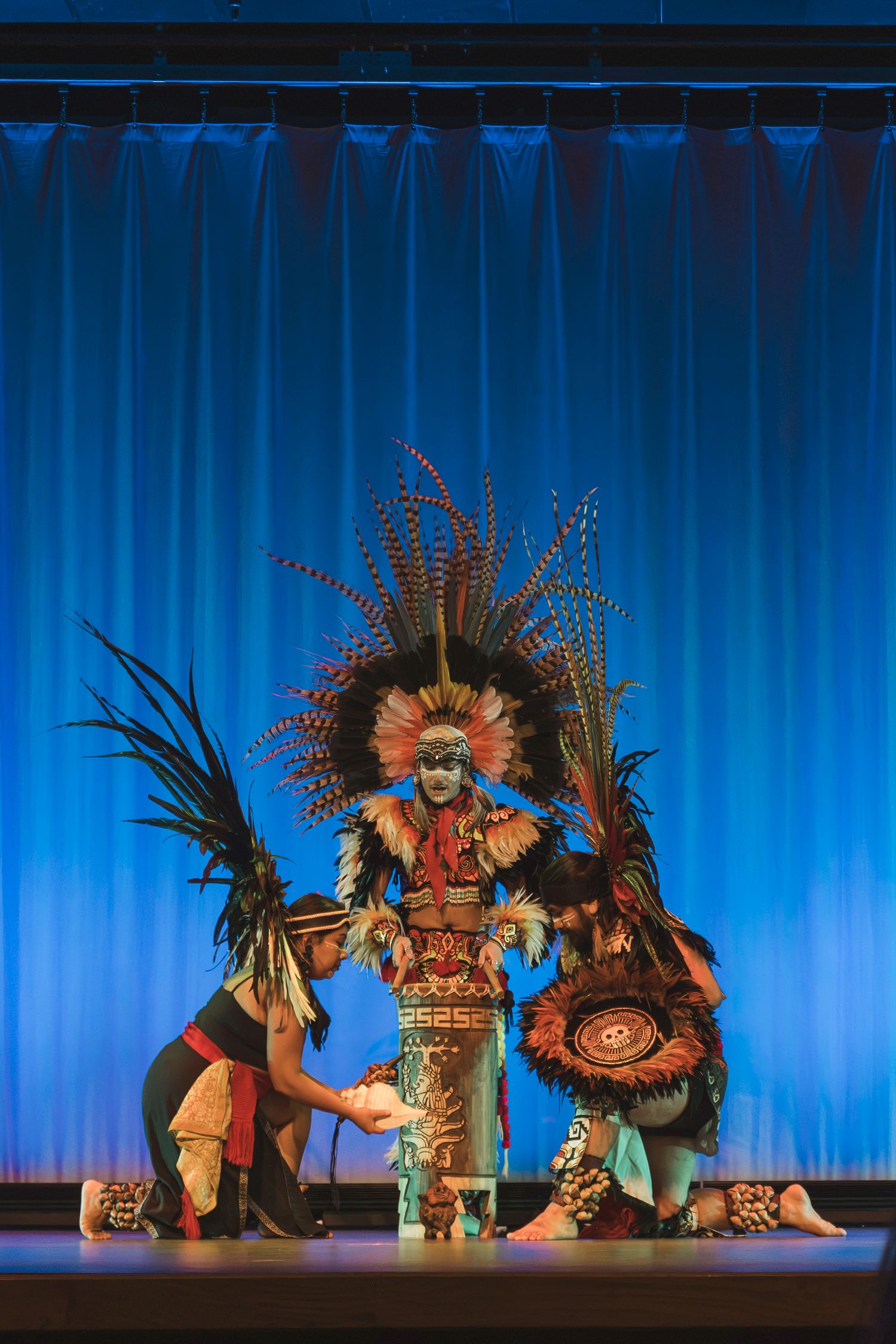 Three performers in elaborate Aztec ceremonial costumes with feathered headdresses on a blue-curtained stage.