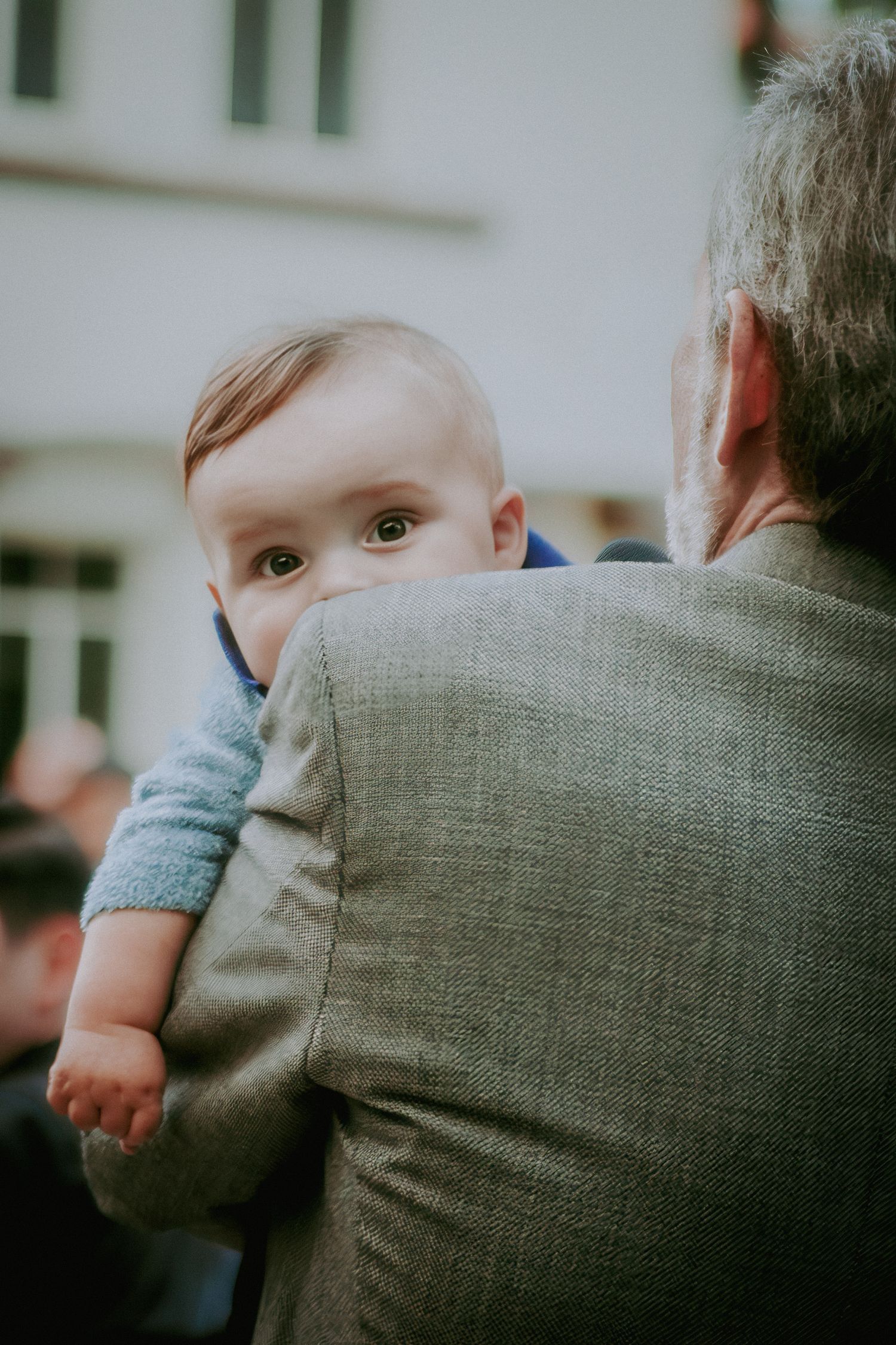 An elderly man in a gray suit holds a wide-eyed baby in a blue outfit, who gazes over his shoulder.