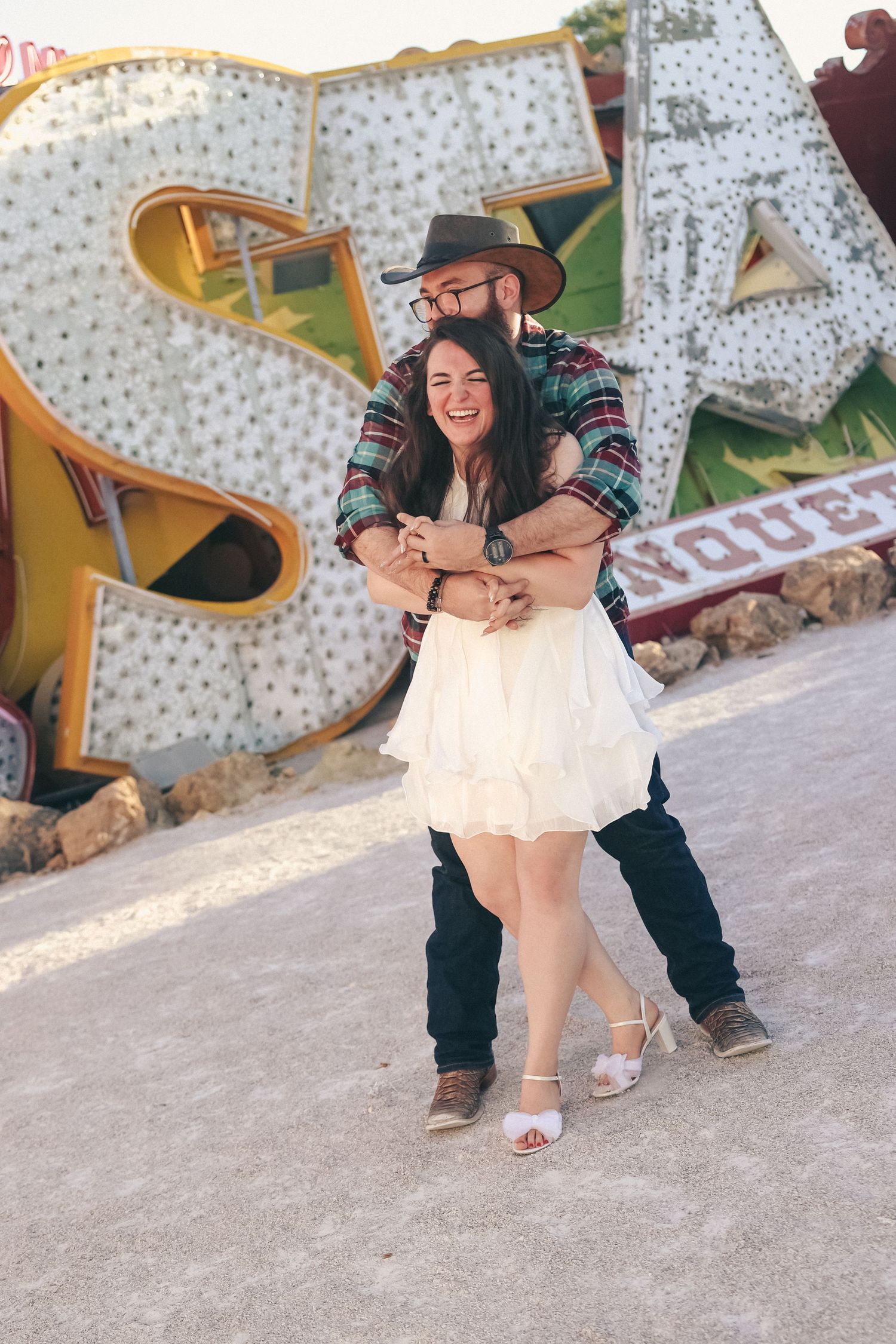 A joyful couple embraces and laughs in front of large vintage neon signs at an outdoor museum.