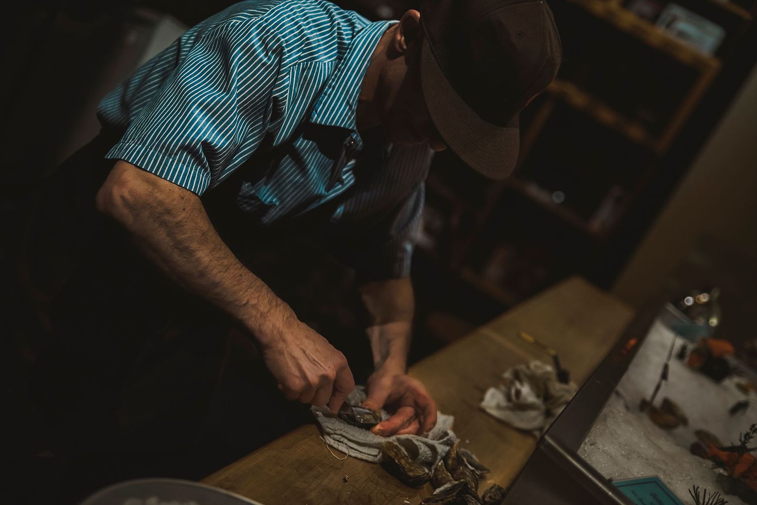 Man in striped shirt and hat shucking oysters on a wooden cutting board in a dimly lit kitchen.