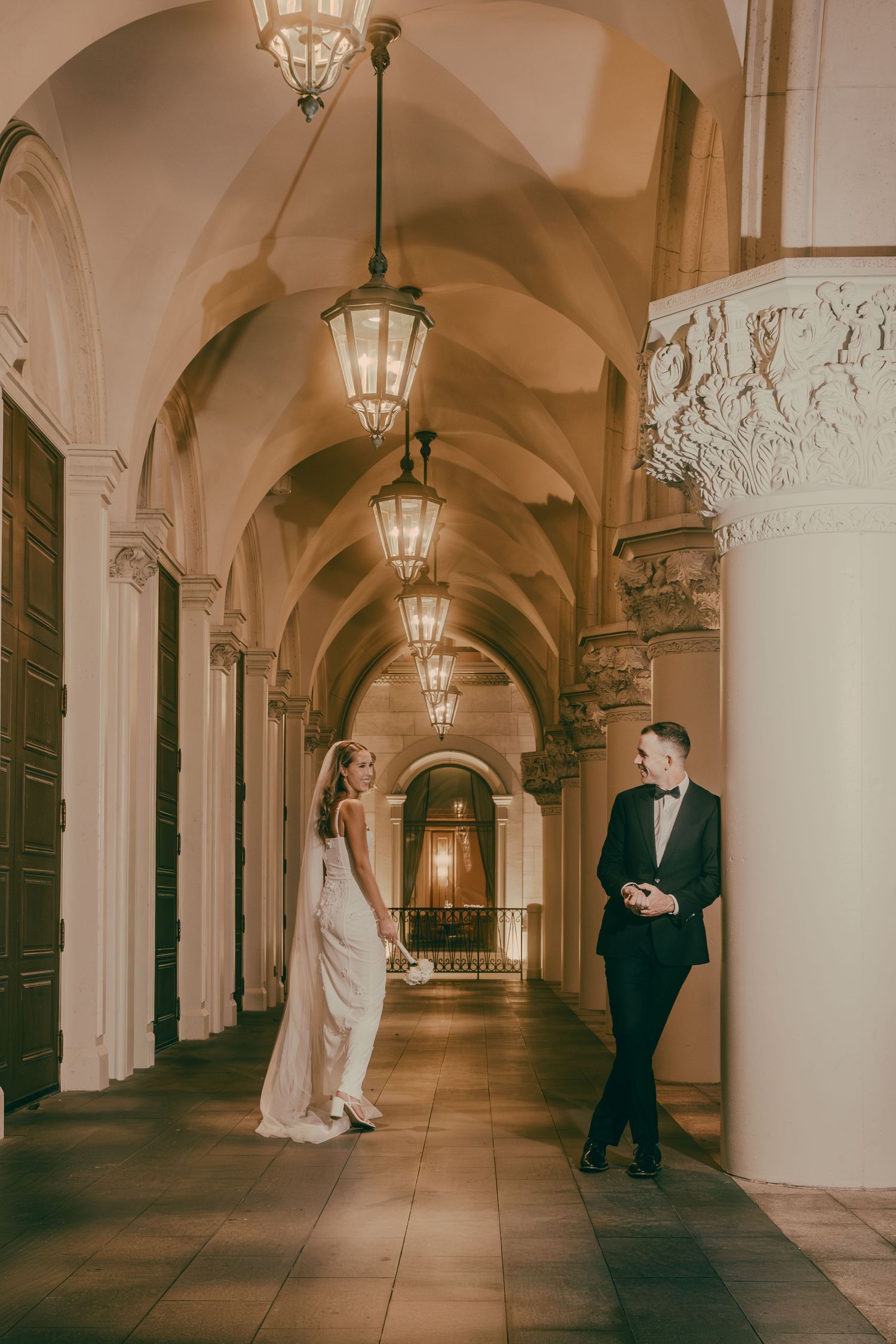 Wedding couple share a romantic moment in a grand illuminated hallway with vaulted ceilings and ornate columns.