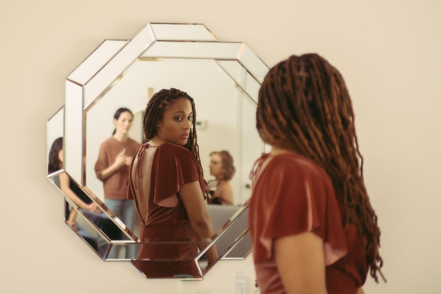 A woman with long locs examines her reflection in an ornate octagonal wall mirror.