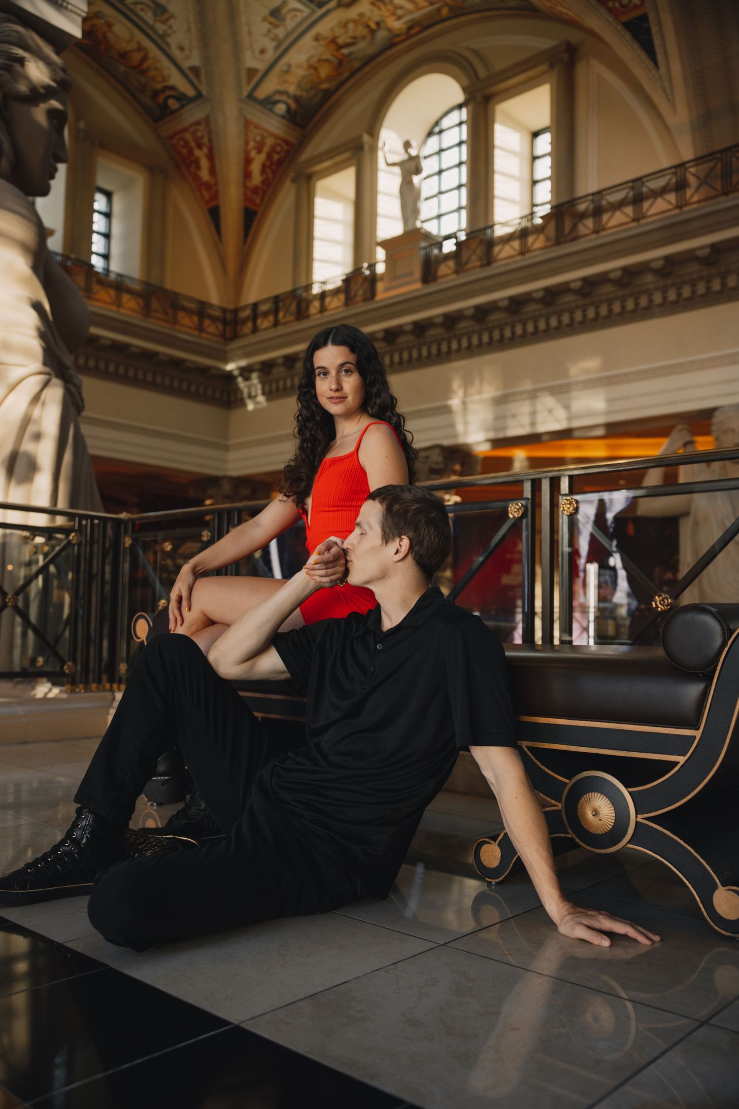 Couple posing in ornate grand hall, woman in red dress seated above man in black on reflective floor.