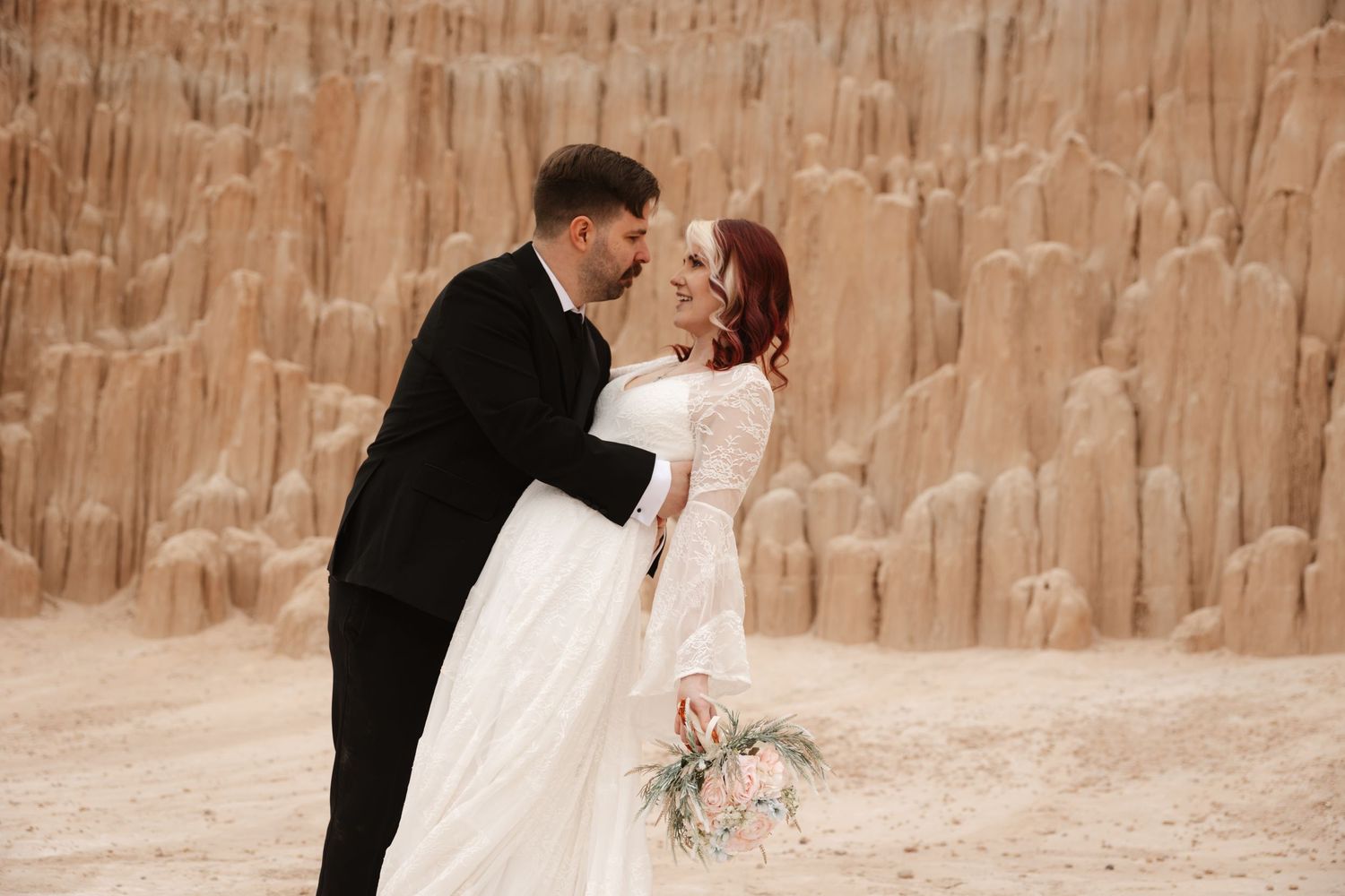 Groom leans in to kiss red-haired bride in white lace dress against towering desert sandstone rock formations.