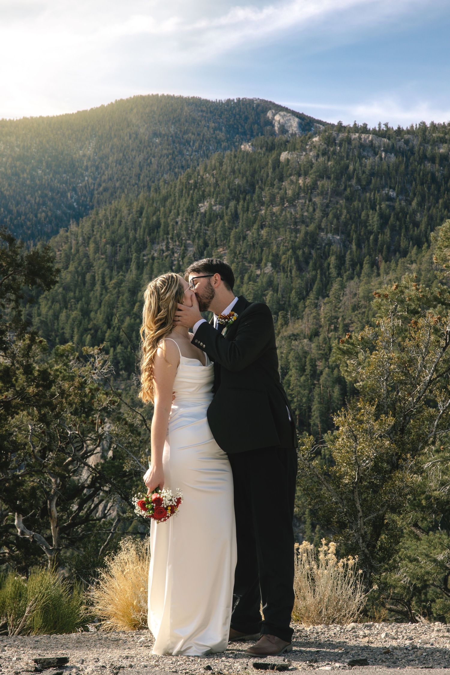 Bride and groom share a kiss with stunning mountain forest backdrop in outdoor wedding photo.