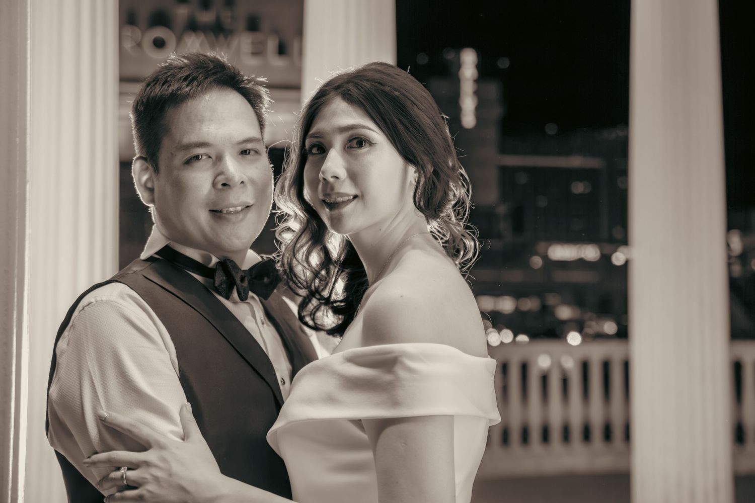 Black and white wedding photo of a smiling couple embracing indoors with city lights glowing in the background at night.