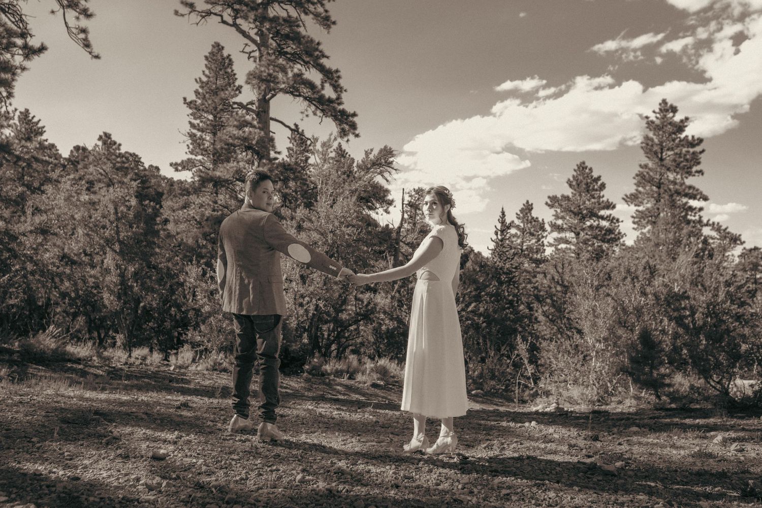 A couple holds hands in a serene forest clearing, captured in a romantic sepia-toned outdoor portrait.