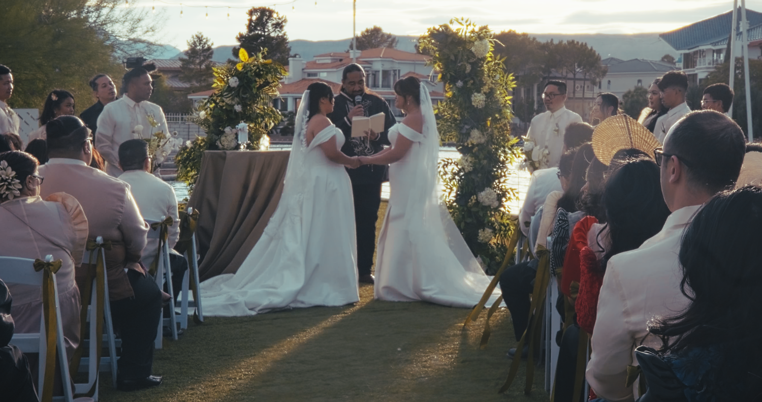 Two brides exchange vows at an outdoor wedding ceremony with guests seated.