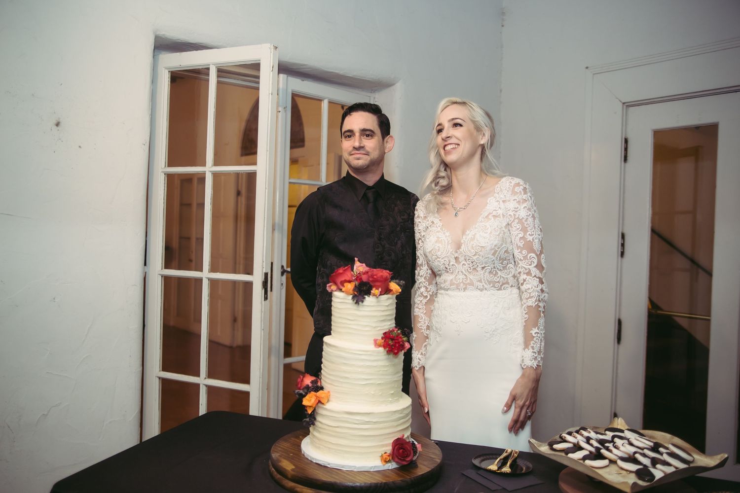 A bride in lace gown and groom in black suit stand smiling beside a three-tier white wedding cake with floral topper.