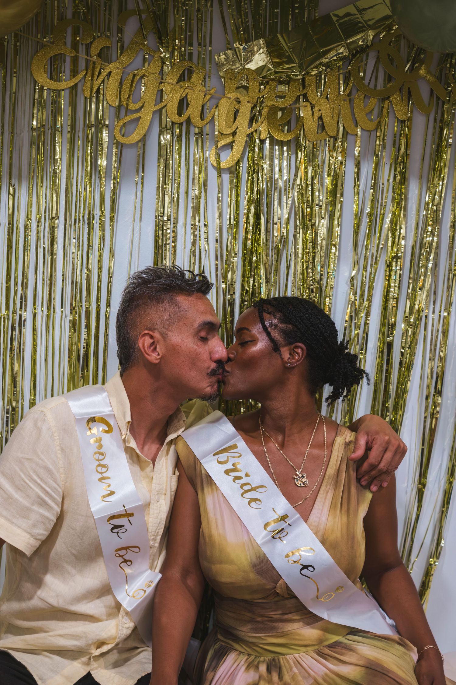 Couple kissing at engagement party with gold tinsel backdrop and Bride and Groom-to-be sashes.