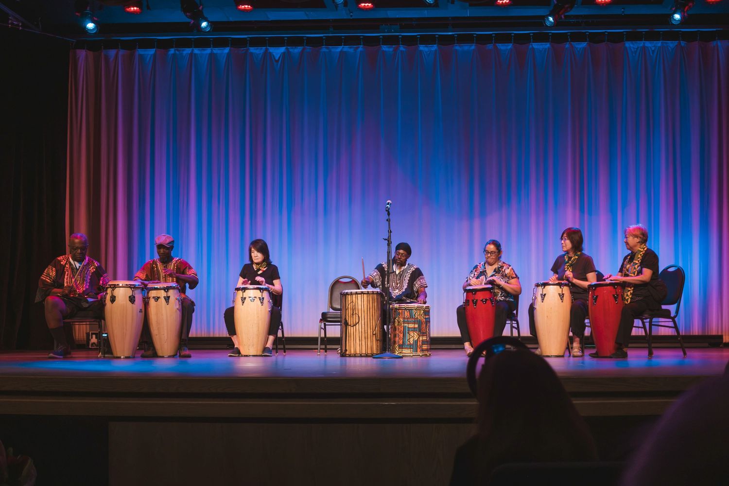 Seven musicians playing conga drums on a colorful stage with blue and pink curtains during a live performance.