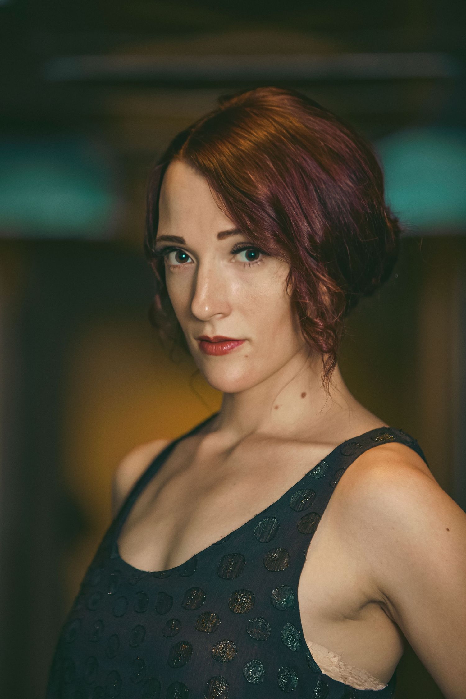 Red-haired woman in black dress posing confidently indoors with bokeh background.
