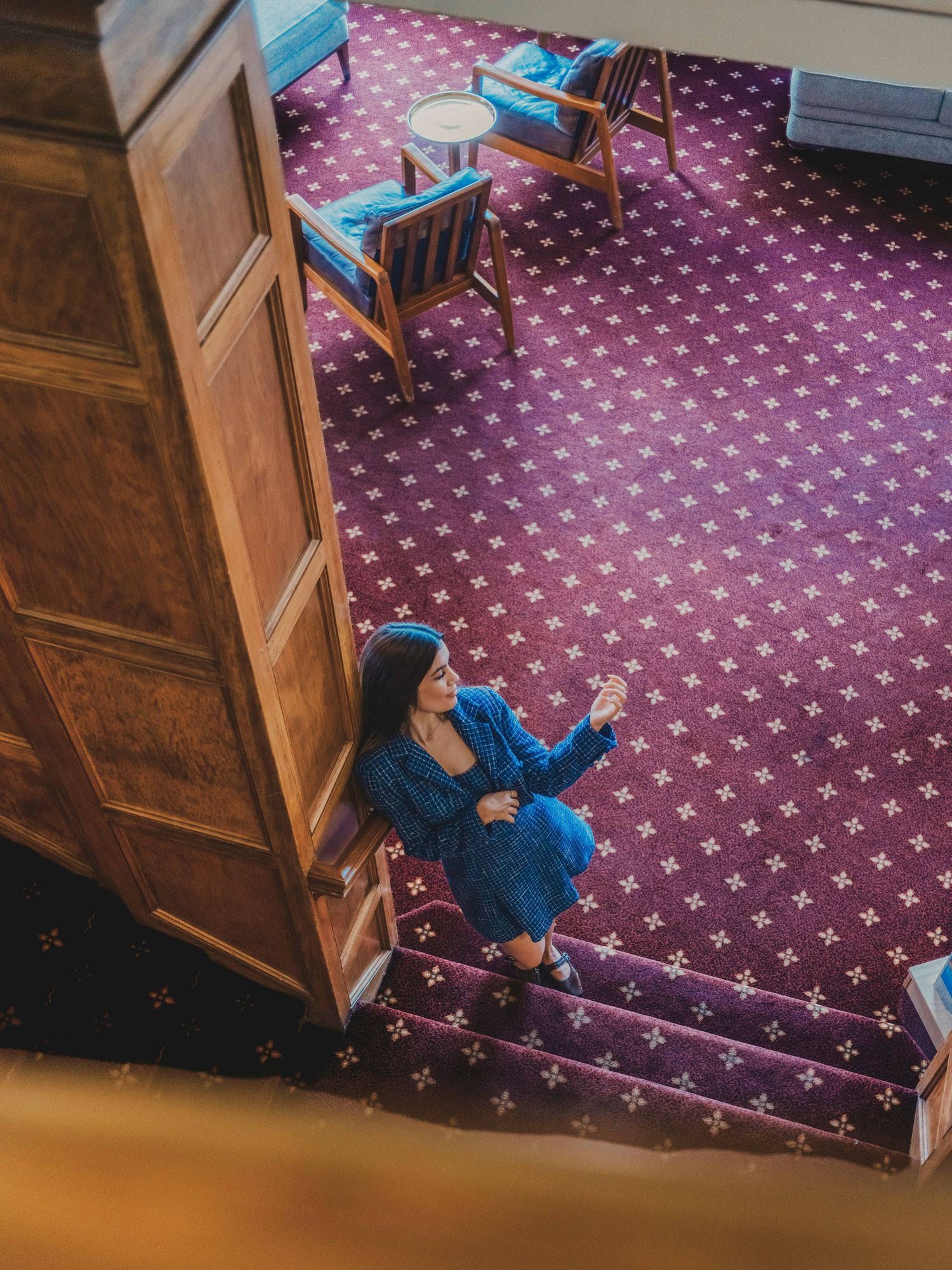 Woman in blue dress sitting on stairs in elegant hotel lobby with purple patterned carpet and wooden furniture.