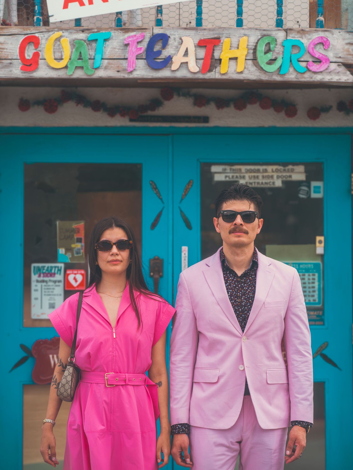 Couple in matching pink outfits posing before colorful Goat Feathers storefront with teal doors.