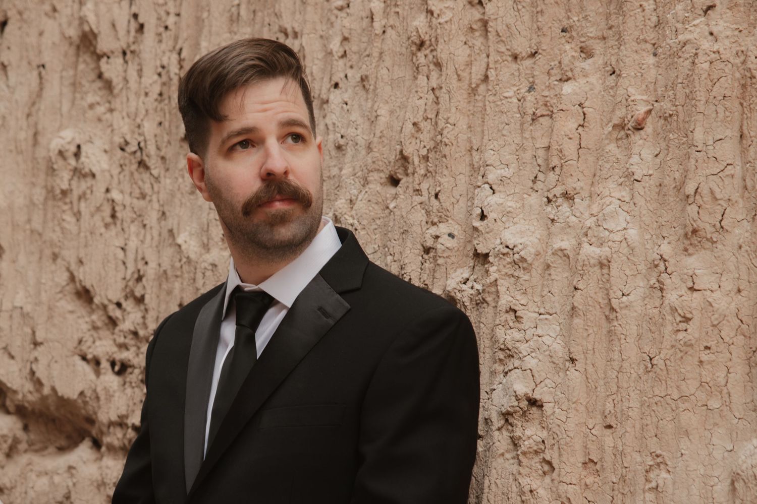 Groom in a black suit and tie with a mustache posing against a sandy textured wall outdoors.